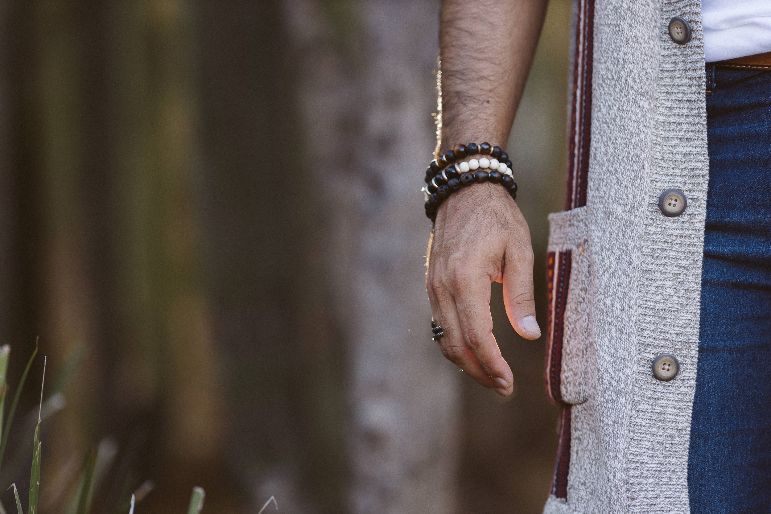 Primer plano de una mano masculina con pulseras de cuentas negras y blancas, al lado de un suéter gris con botones grandes.