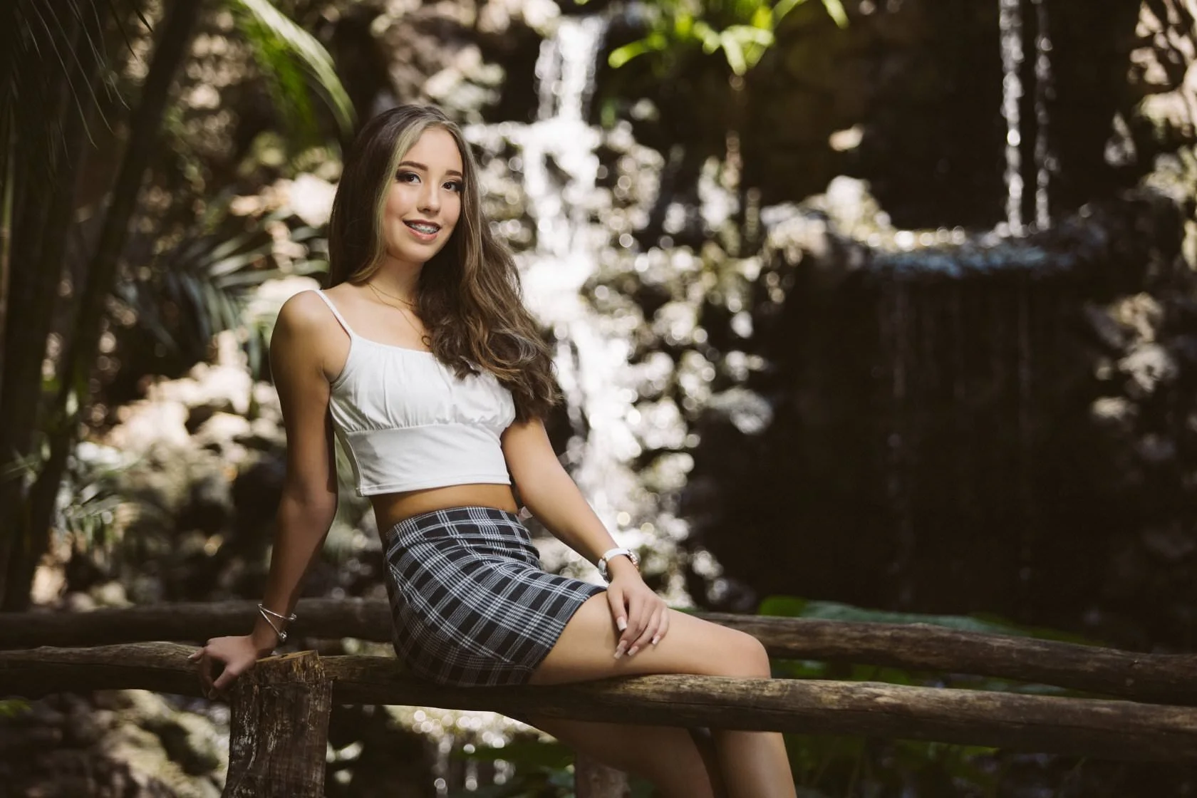 Mujer joven sentada en una cerca de madera frente a una cascada, vistiendo una blusa blanca y una falda a cuadros.