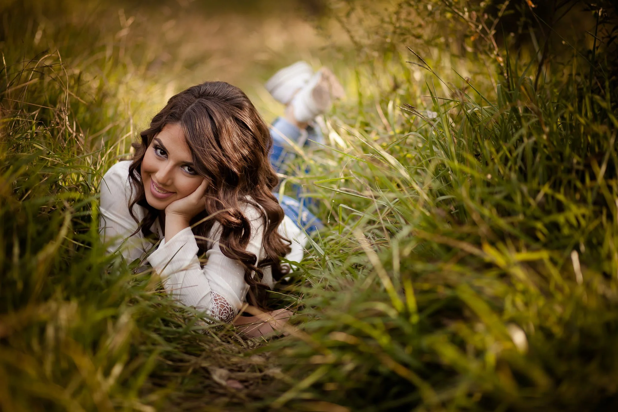 Mujer sonriendo acostada en el pasto, vestida con blusa blanca y jeans, con cabello suelto.