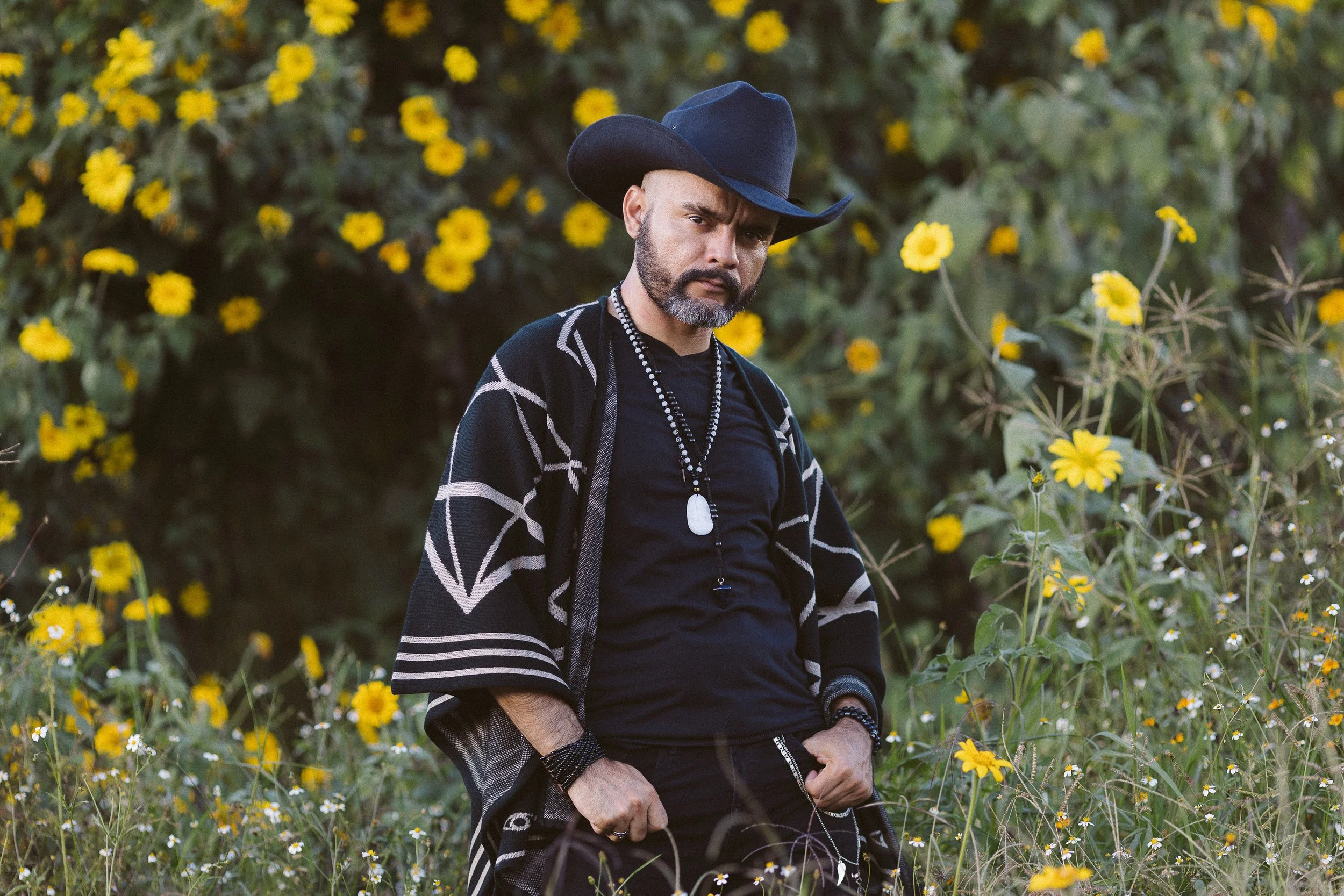 Hombre con sombrero negro y poncho en campo con flores amarillas.