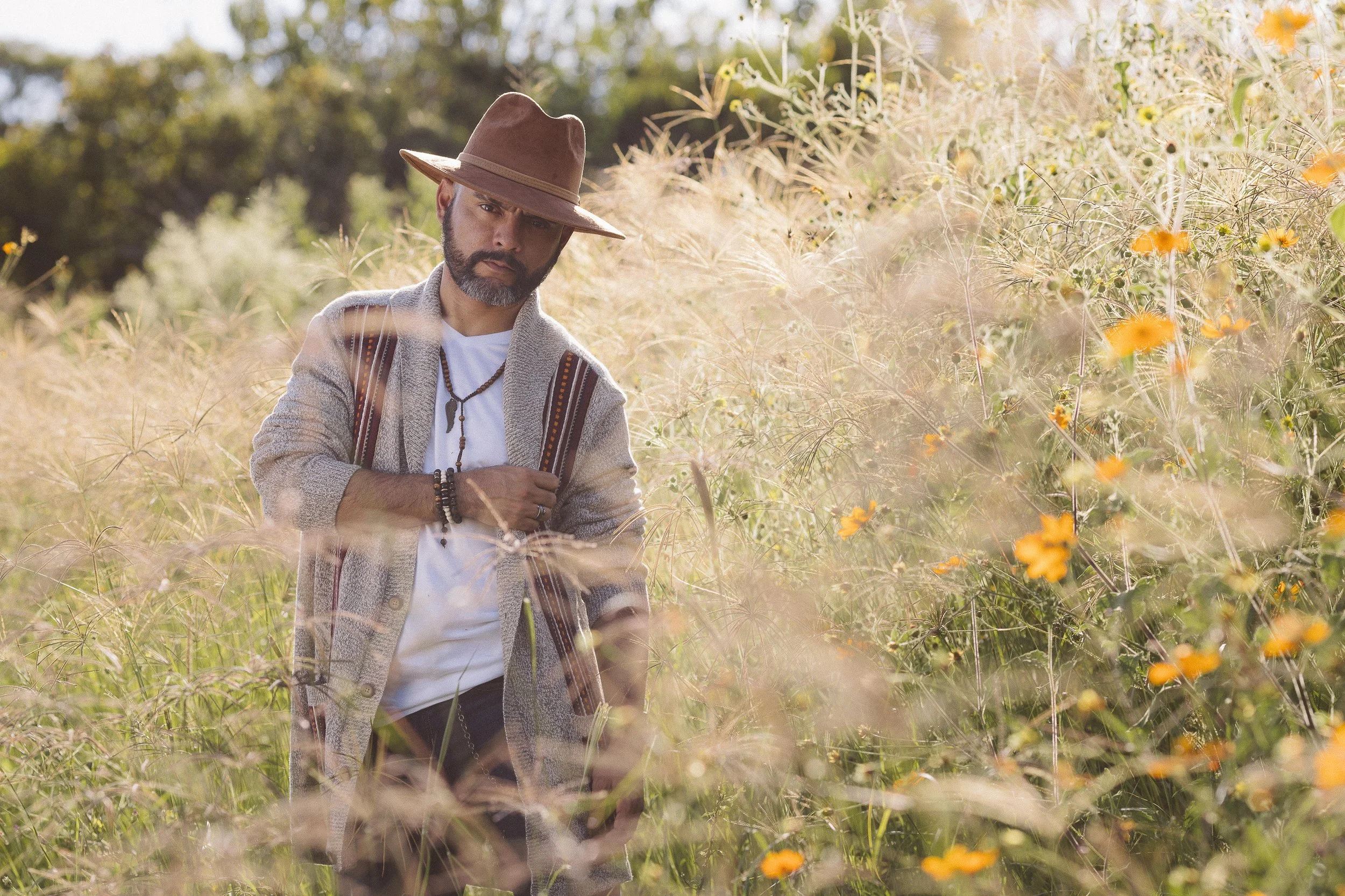 Hombre con sombrero marrón y suéter caminando en campo con flores amarillas y vegetación verde.