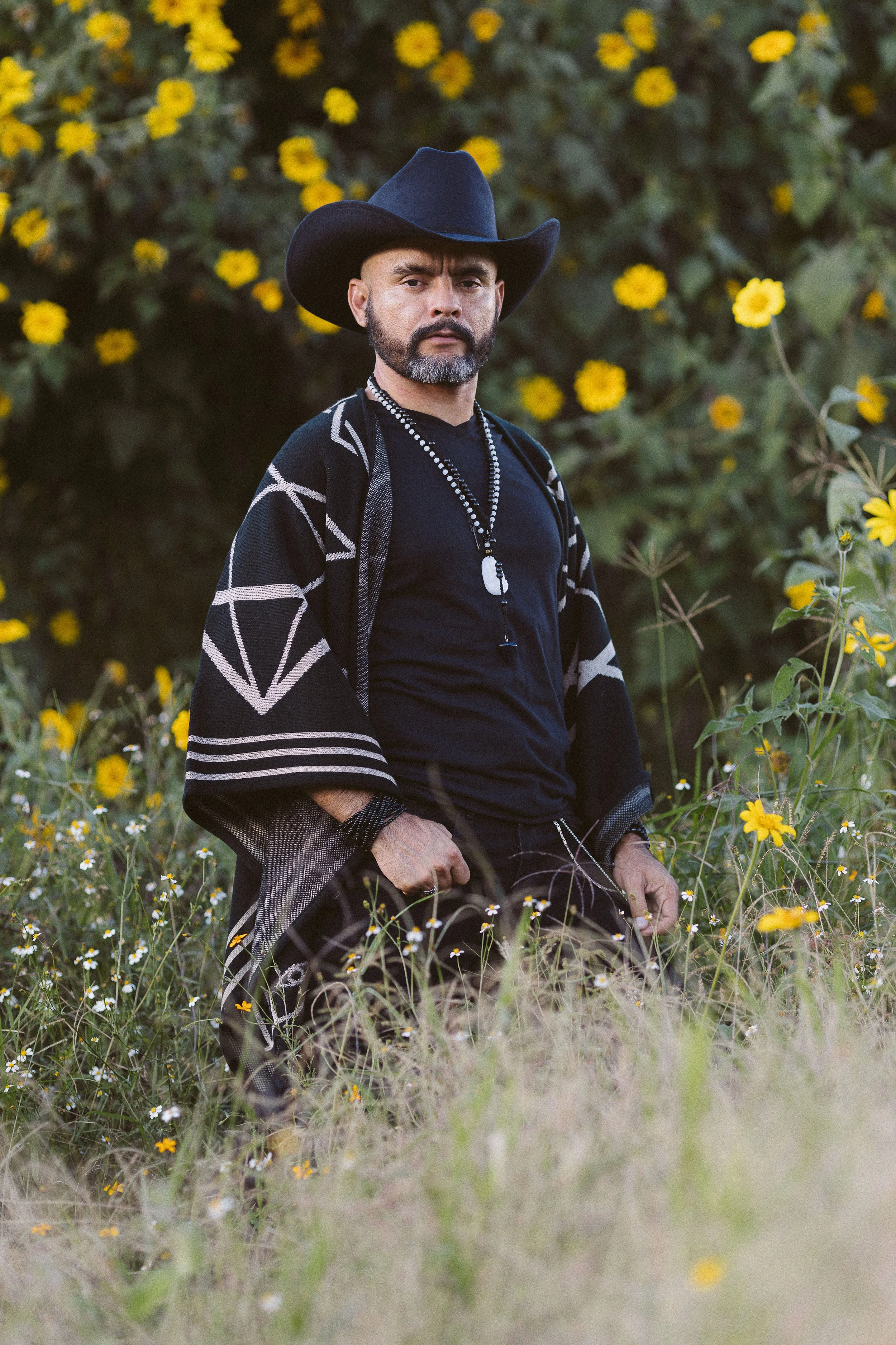 Hombre con sombrero de vaquero y poncho, rodeado de flores amarillas en un campo.
