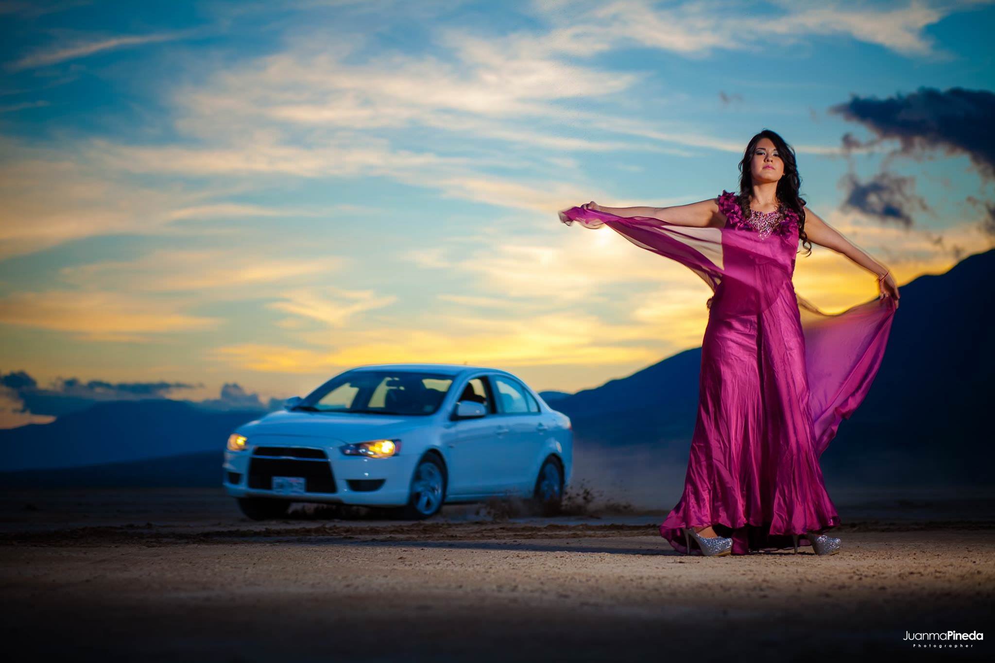 Mujer con vestido rosa posando en un paisaje desértico al atardecer, con un auto blanco en movimiento en el fondo.