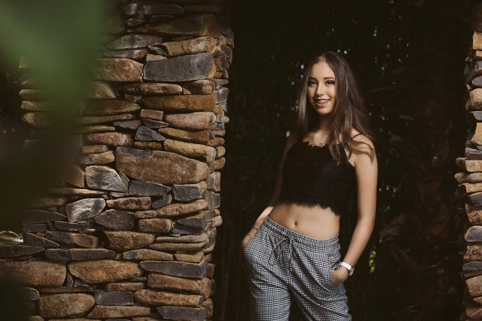 Joven sonriendo de pie entre pilares de piedra, con top negro y pantalones a cuadros.