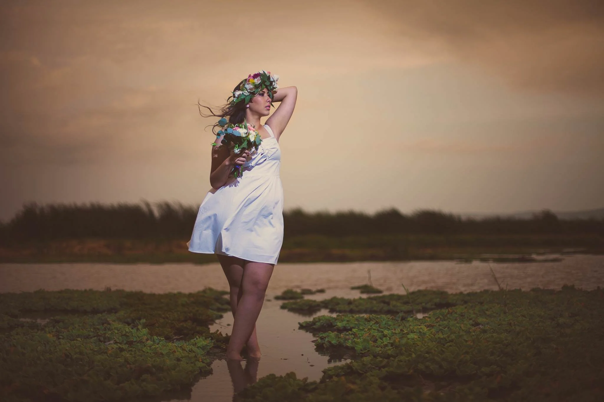 Mujer con vestido blanco y corona de flores, sosteniendo un ramo, parada en un estanque con plantas acuáticas.