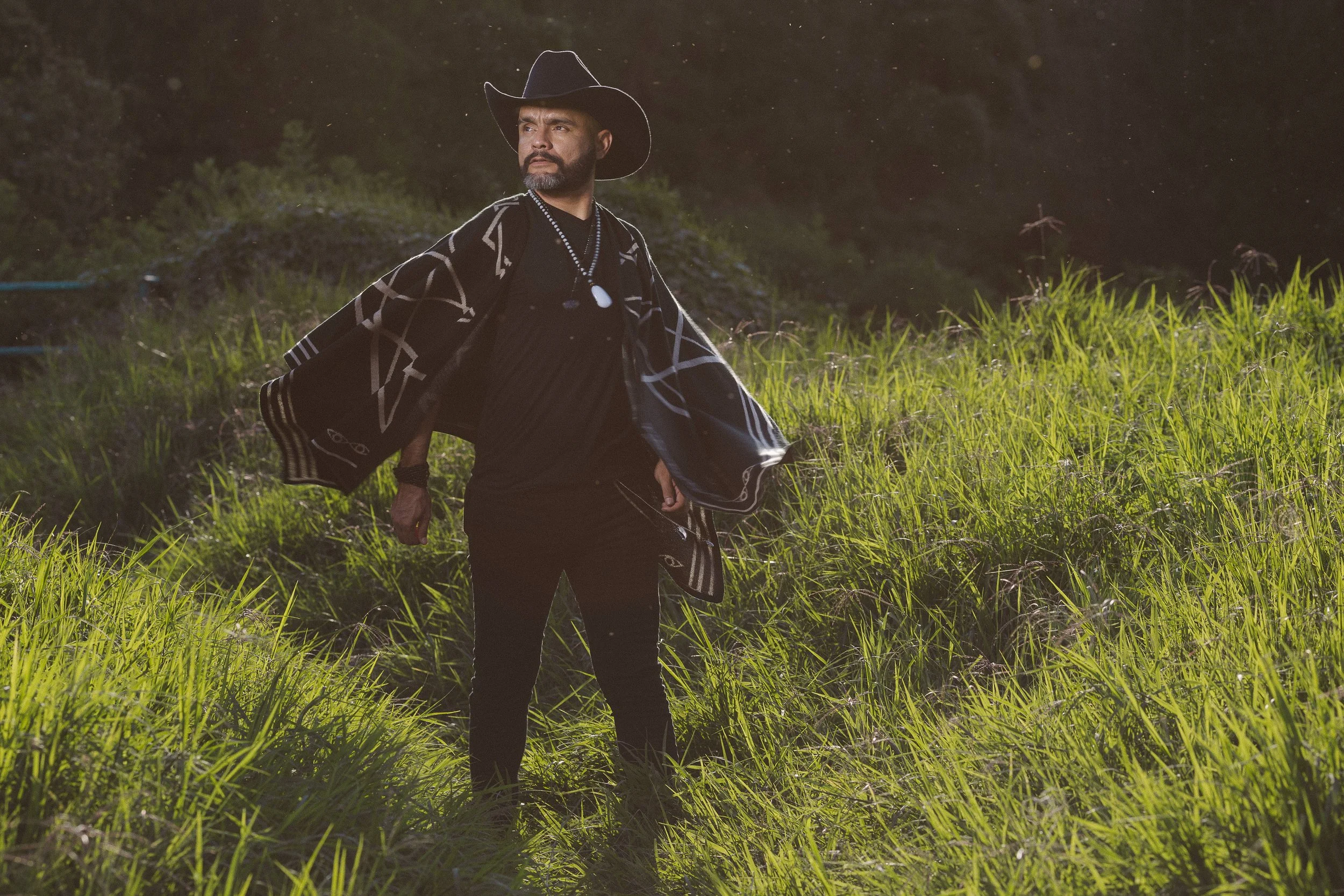 Hombre con sombrero vaquero y poncho negro en campo de hierba verde.