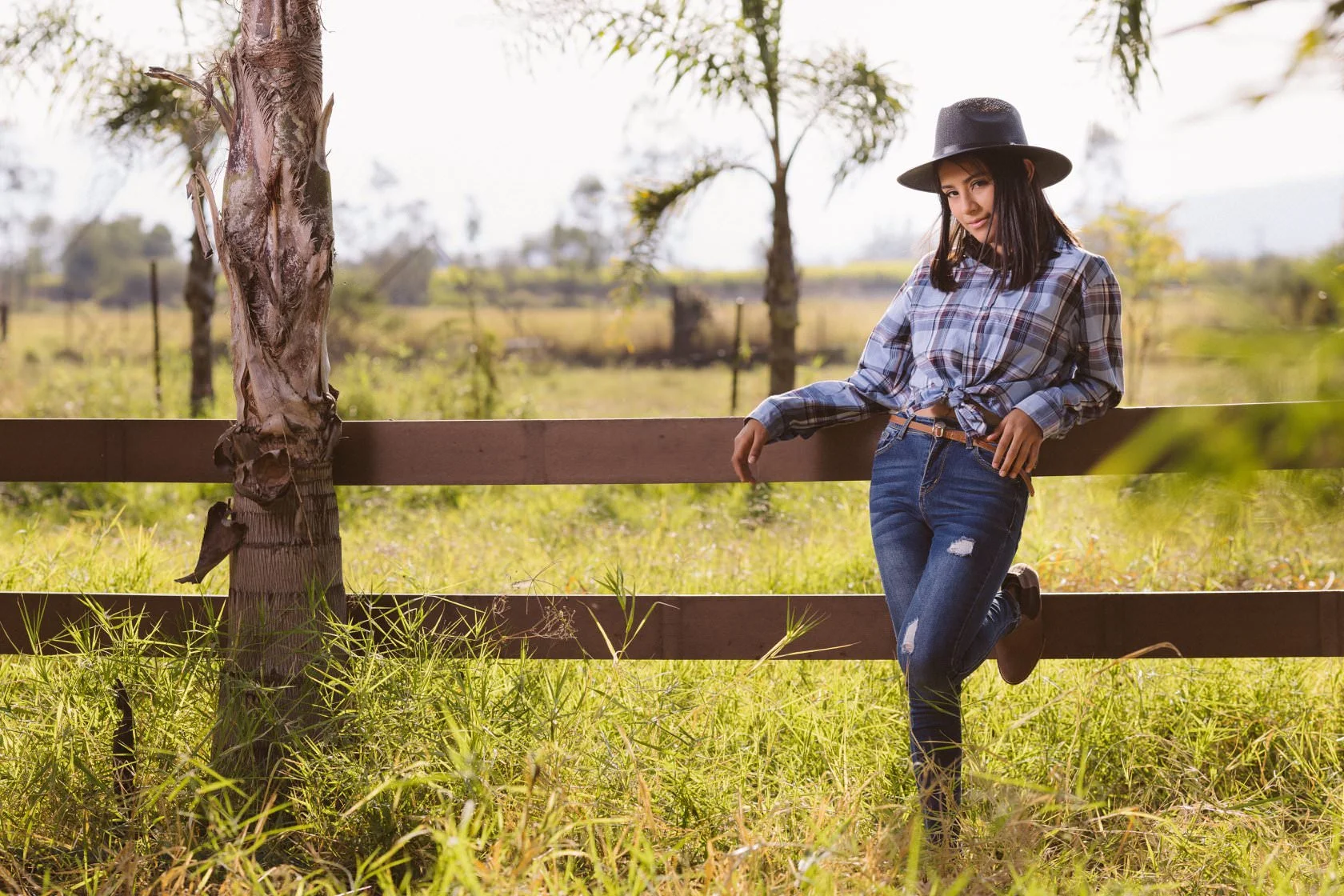 Mujer joven con sombrero y camisa a cuadros posando junto a una cerca en un campo soleado con palmeras.