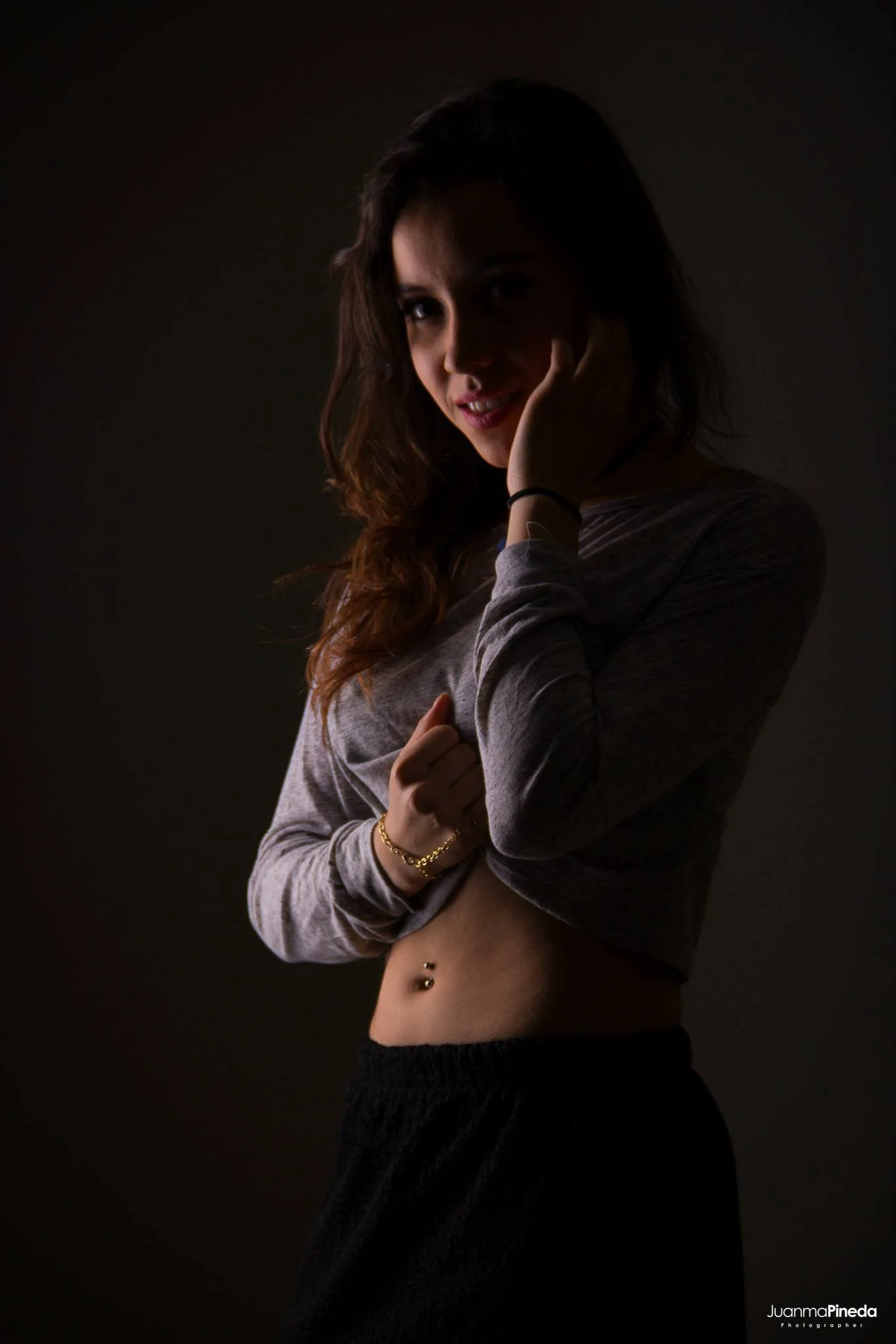 Mujer joven con cabello suelto y largo, usando una blusa gris de manga larga, fotografiada contra un fondo oscuro. Lleva una pulsera dorada y un piercing en el ombligo.