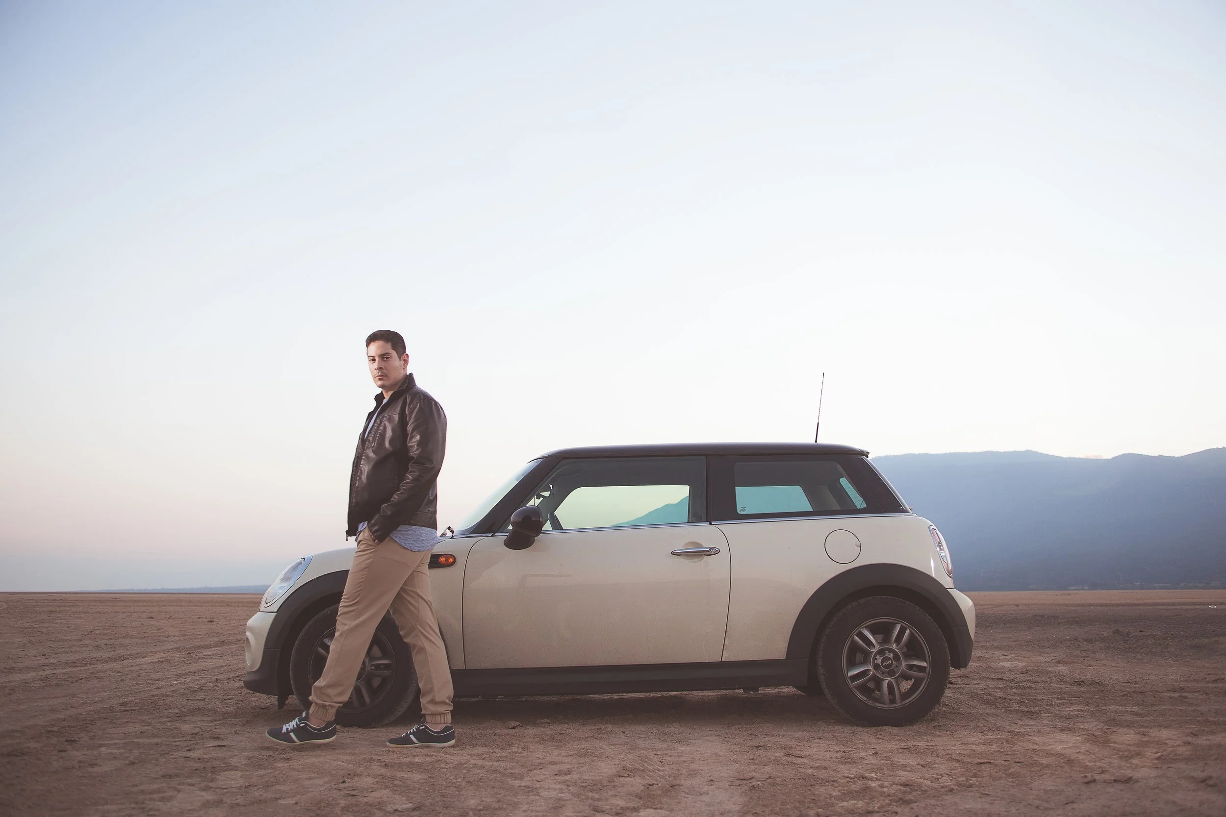 Hombre de pie junto a un auto Mini Cooper blanco en un paisaje desértico.