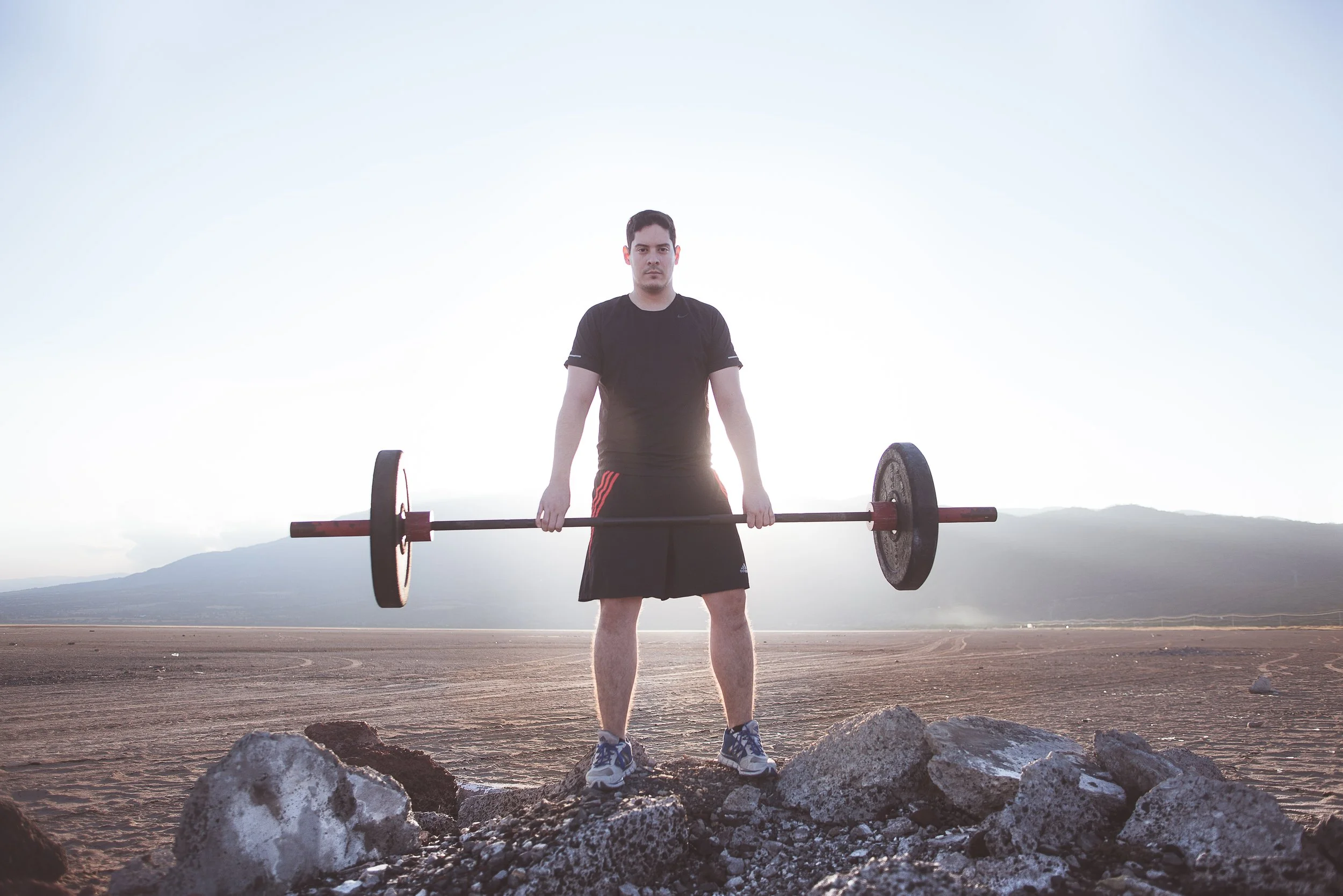 Hombre levantando barra con pesas al aire libre, en un paisaje árido y montañoso.
