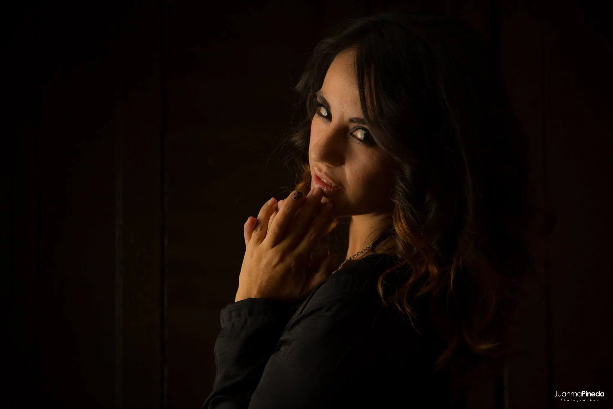 Mujer de cabello rizado y oscuro con expresión pensativa y manos juntas en un fondo oscuro.