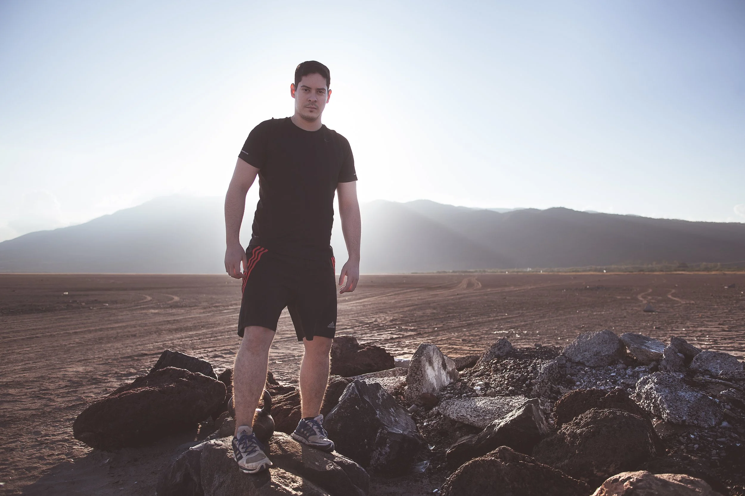 Hombre de pie sobre rocas en un paisaje desértico con montañas al fondo y luz del sol