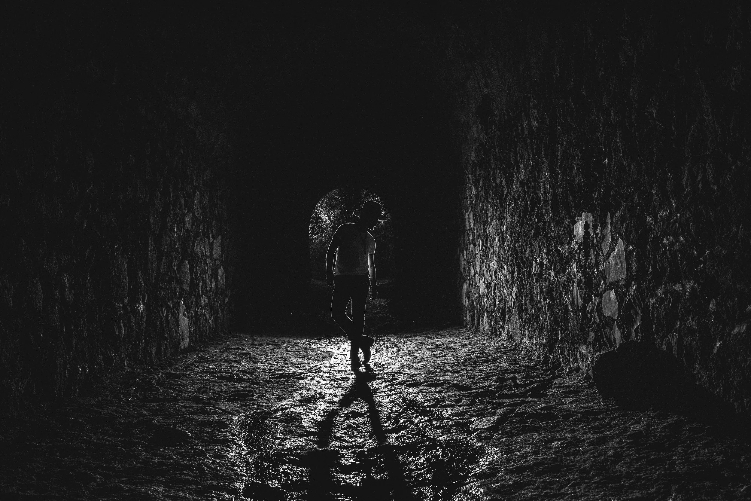 Silueta de una persona caminando en un túnel oscuro con paredes de piedra, iluminado desde atrás, creando sombras en el suelo.