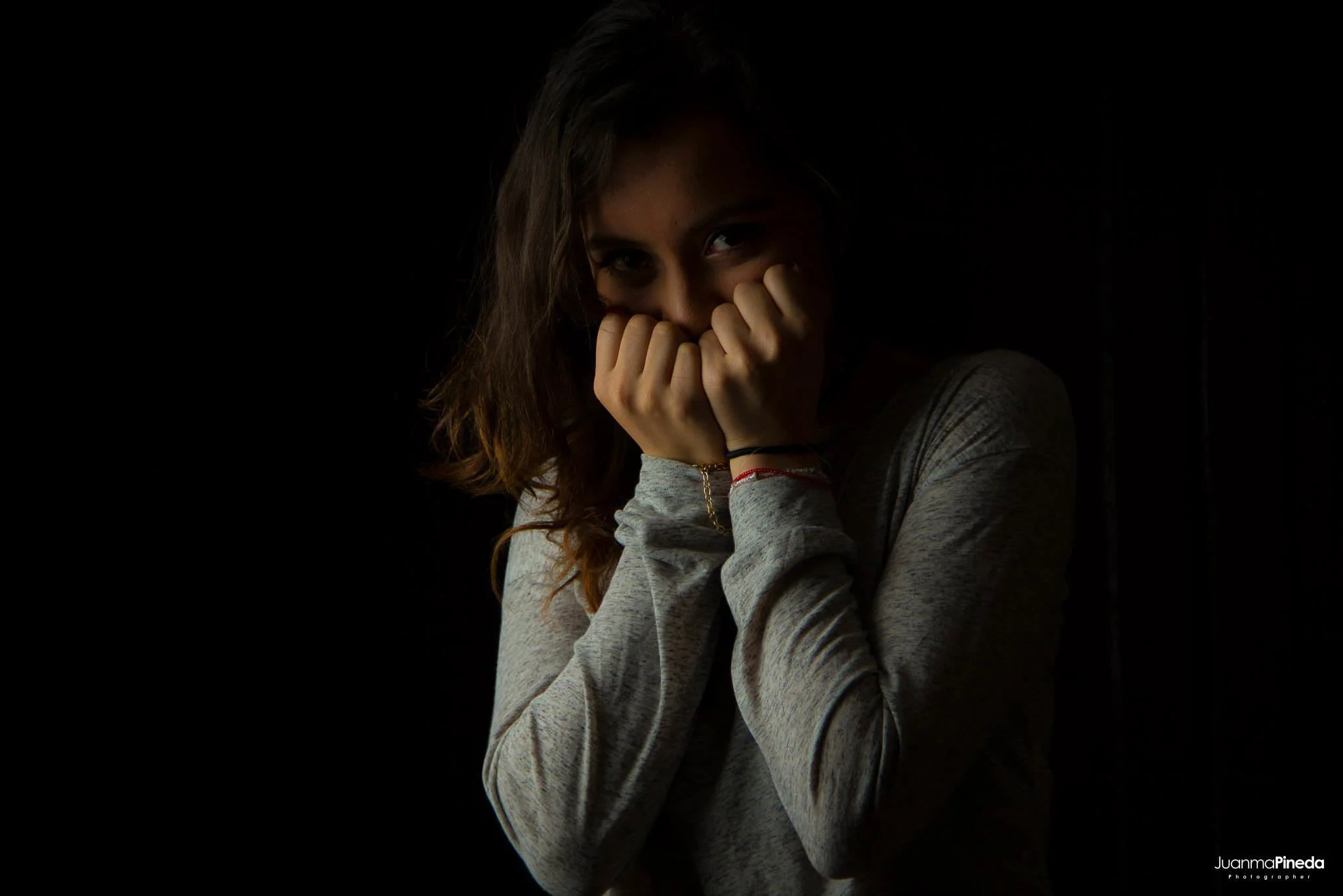 Mujer con cabello suelto, cubriendo su rostro parcialmente con las manos, en un fondo oscuro.