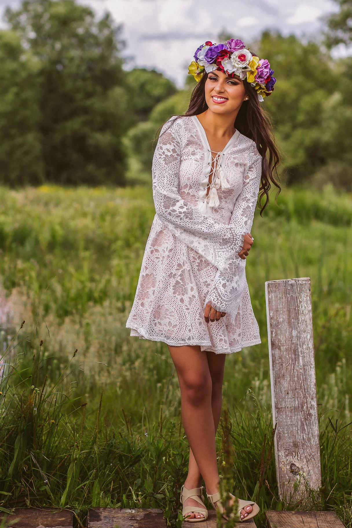 Mujer con vestido blanco de encaje y corona de flores en la cabeza, sonriendo en un campo verde.