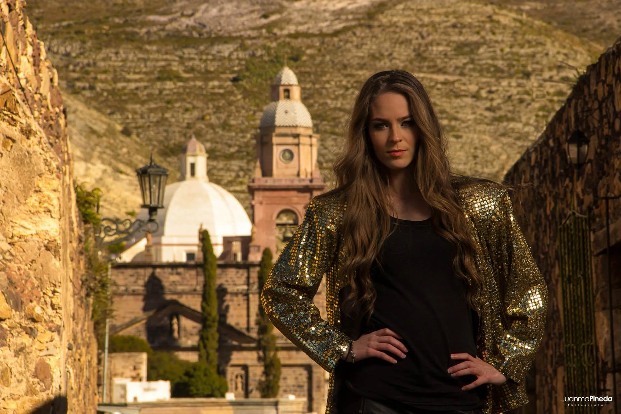 Mujer con chaqueta brillante posando frente a un edificio histórico y paisaje montañoso.