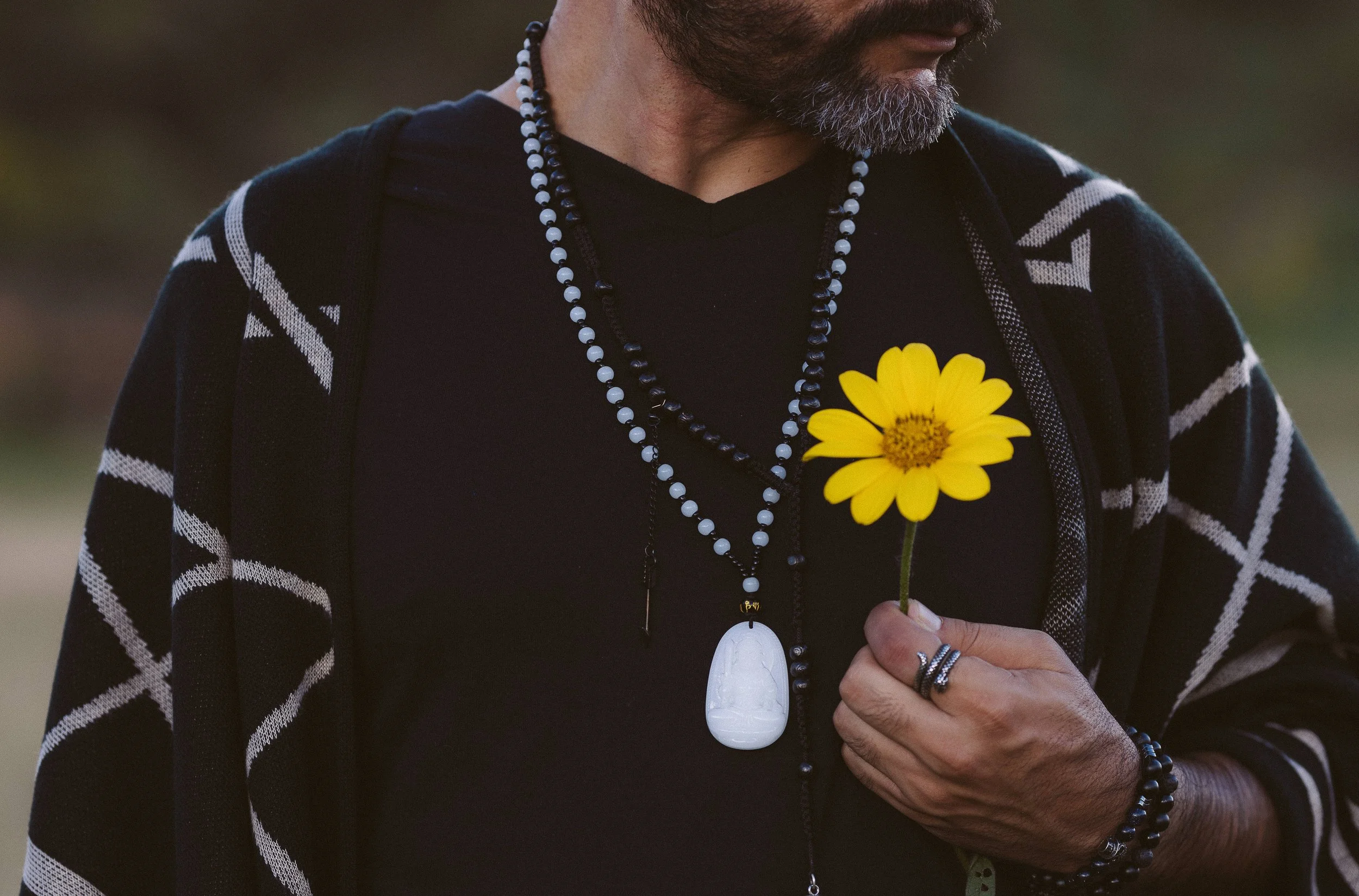 Hombre sosteniendo una flor amarilla, con collares y anillos, vistiendo un suéter con patrón geométrico.