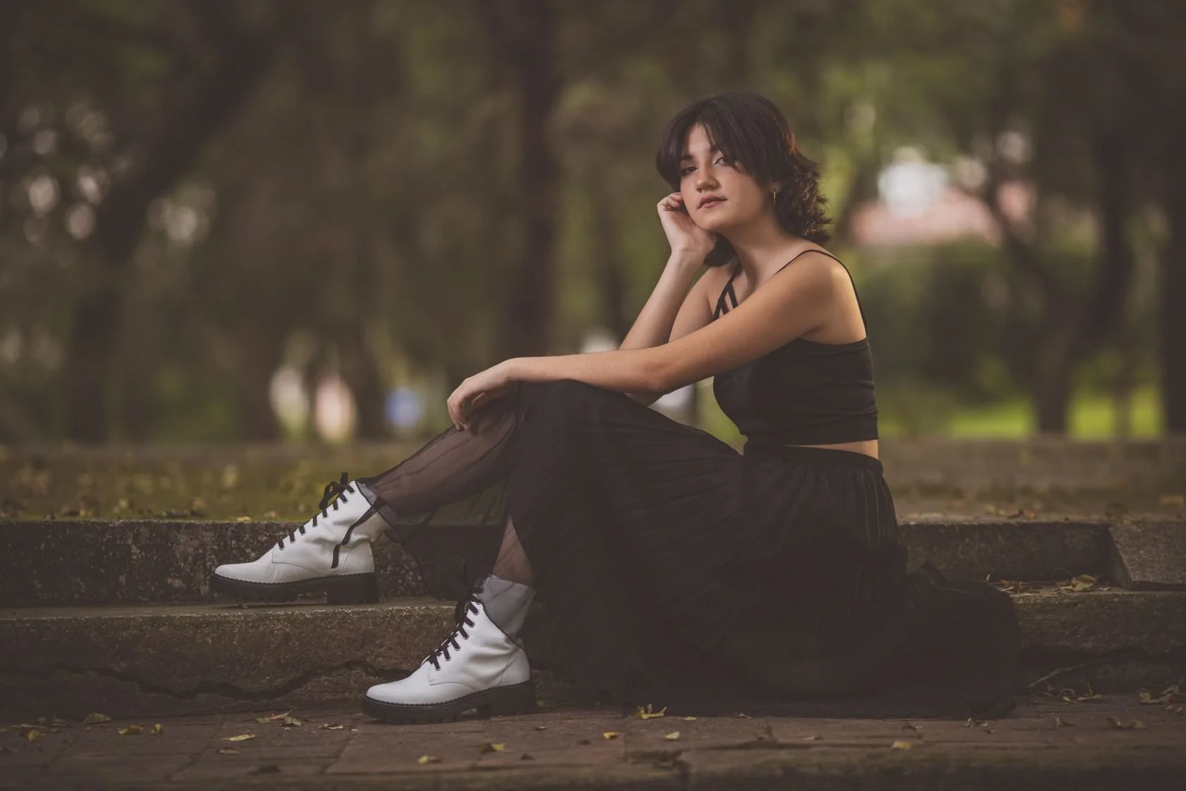 Joven sentada en escaleras al aire libre, vestida de negro con botas blancas, en un entorno natural.