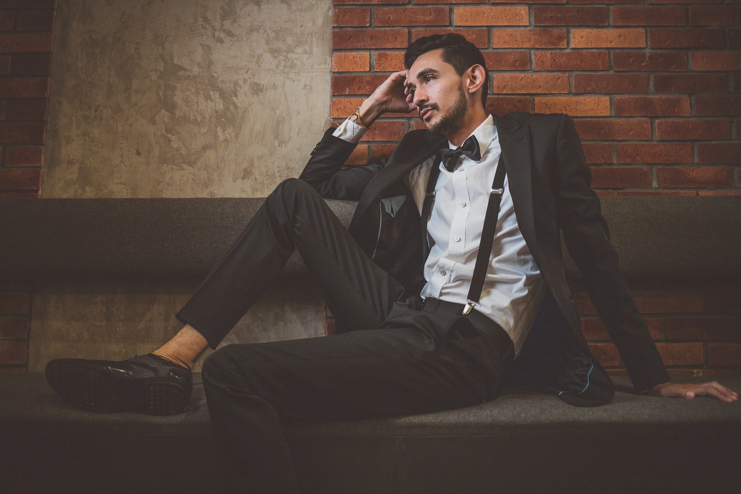 Hombre con esmoquin sentado en un sofá, apoyado contra una pared de ladrillos.