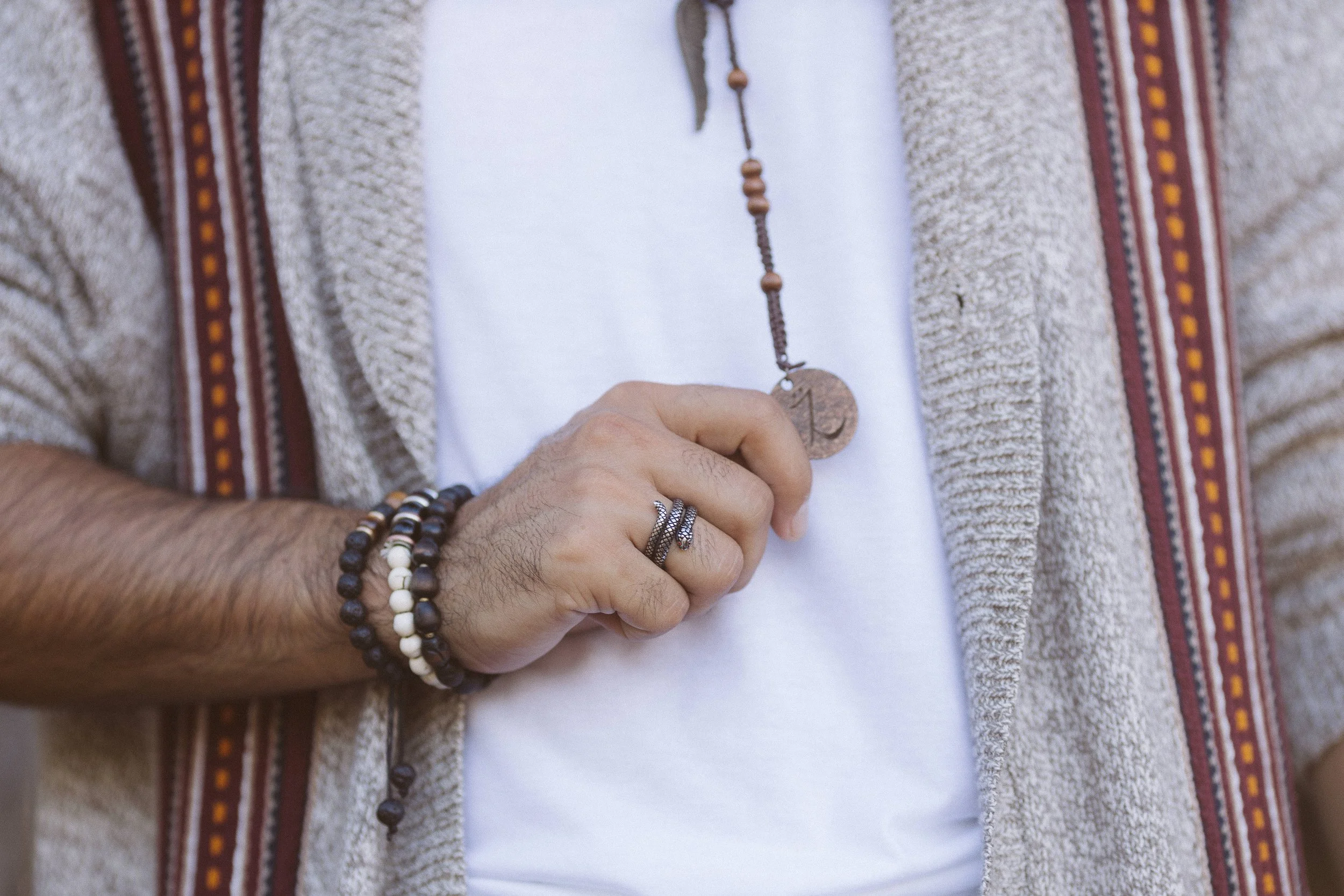 Hombre vistiendo suéter gris, camisa blanca, collares y pulseras. Lleva un colgante y anillos en la mano, mostrando un estilo bohemio.