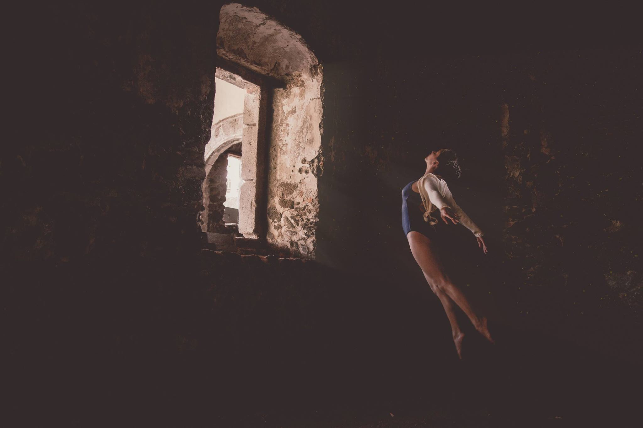 Bailarina en leotardo azul saltando en un ambiente oscuro cerca de una ventana de piedra.