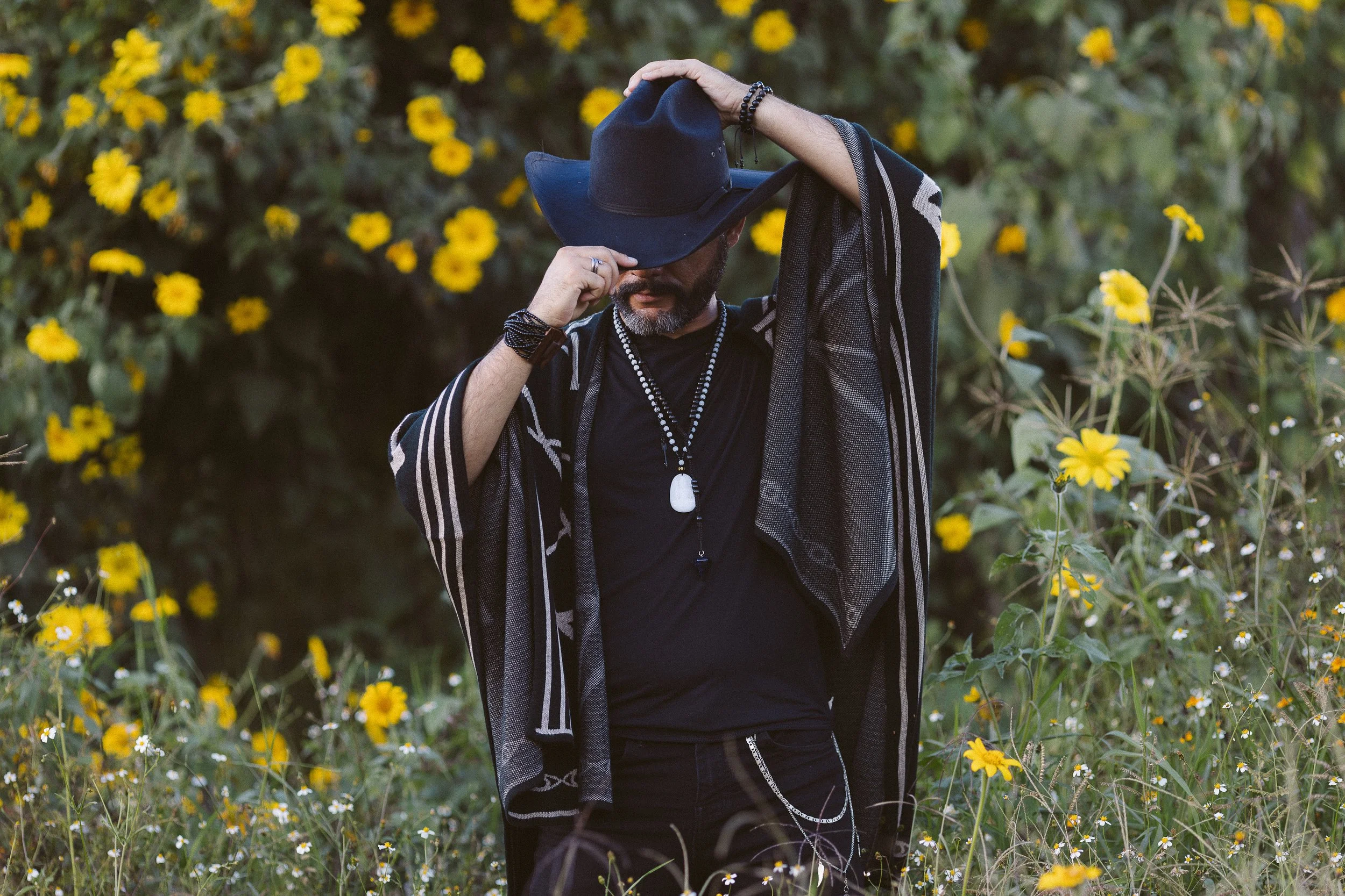 Hombre con sombrero negro, poncho y accesorios, de pie en un campo con flores amarillas.