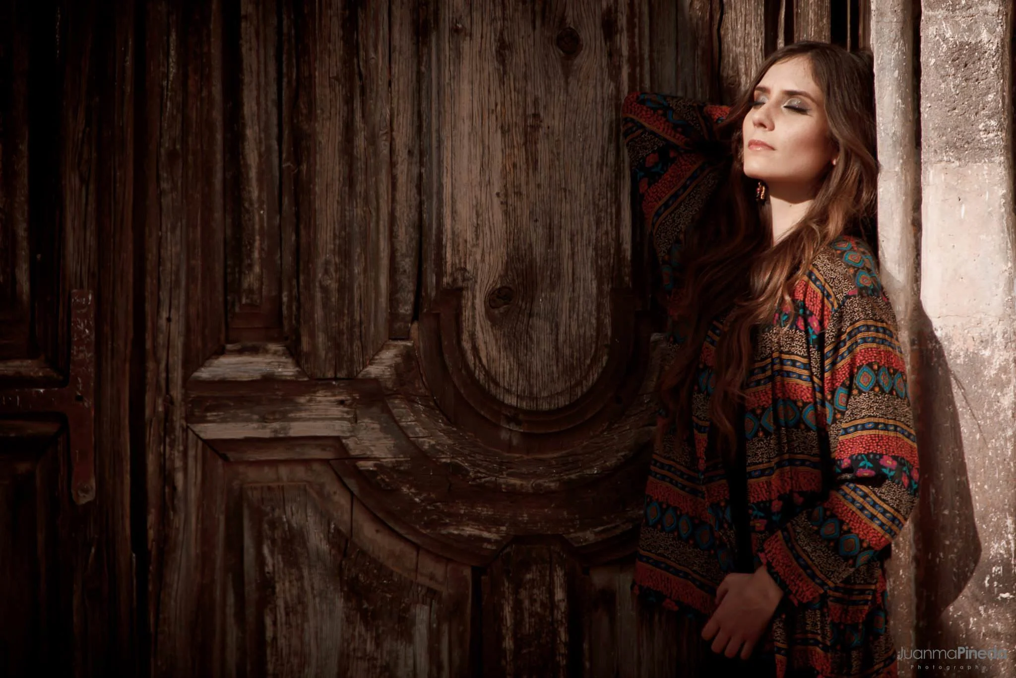 Mujer de cabello largo y suelto frente a una puerta de madera envejecida, lleva un suéter colorido y está con los ojos cerrados.