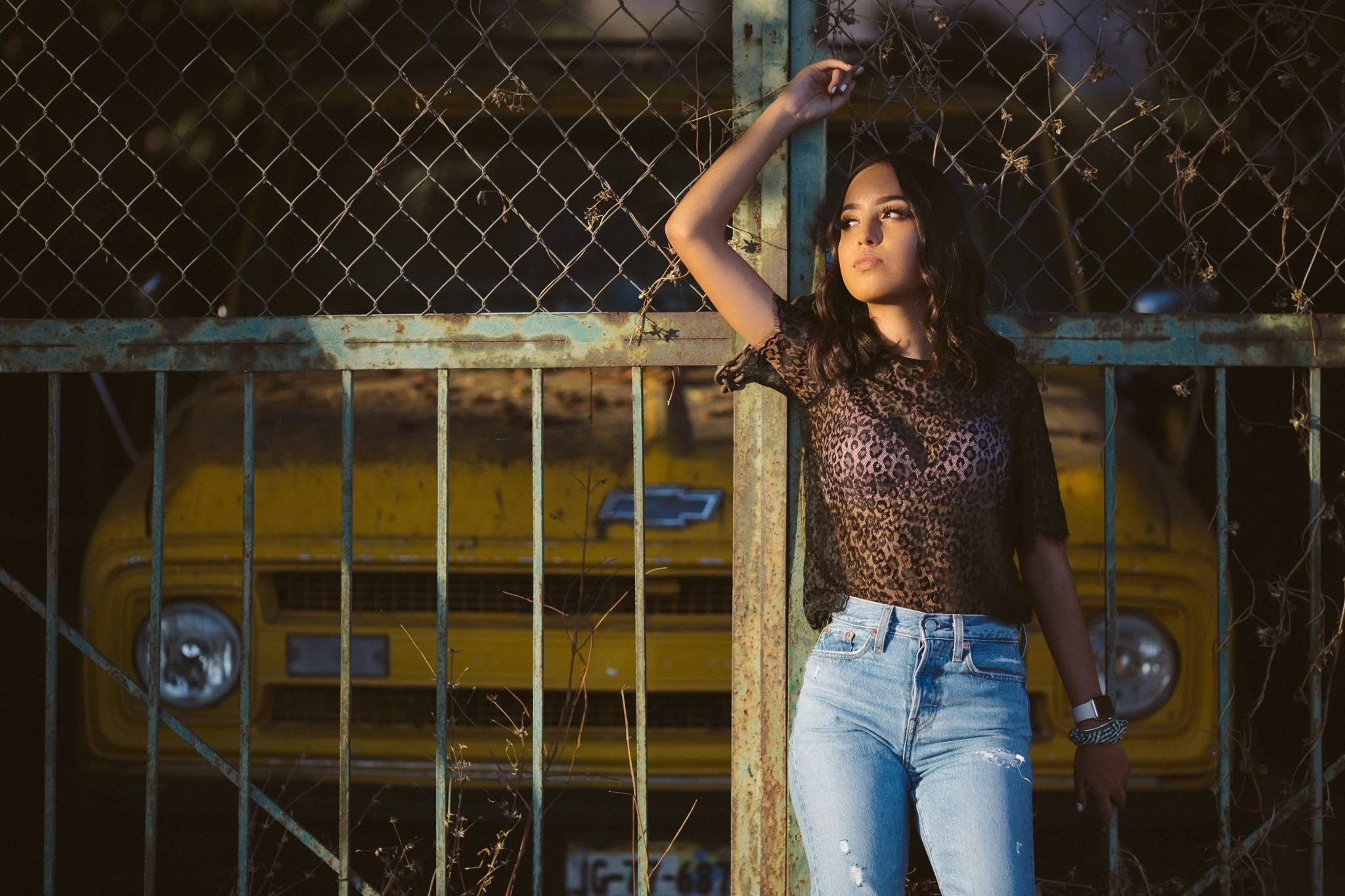 Mujer de pie frente a una reja metálica oxidada, con un auto amarillo de fondo. Lleva una blusa estampada y jeans.