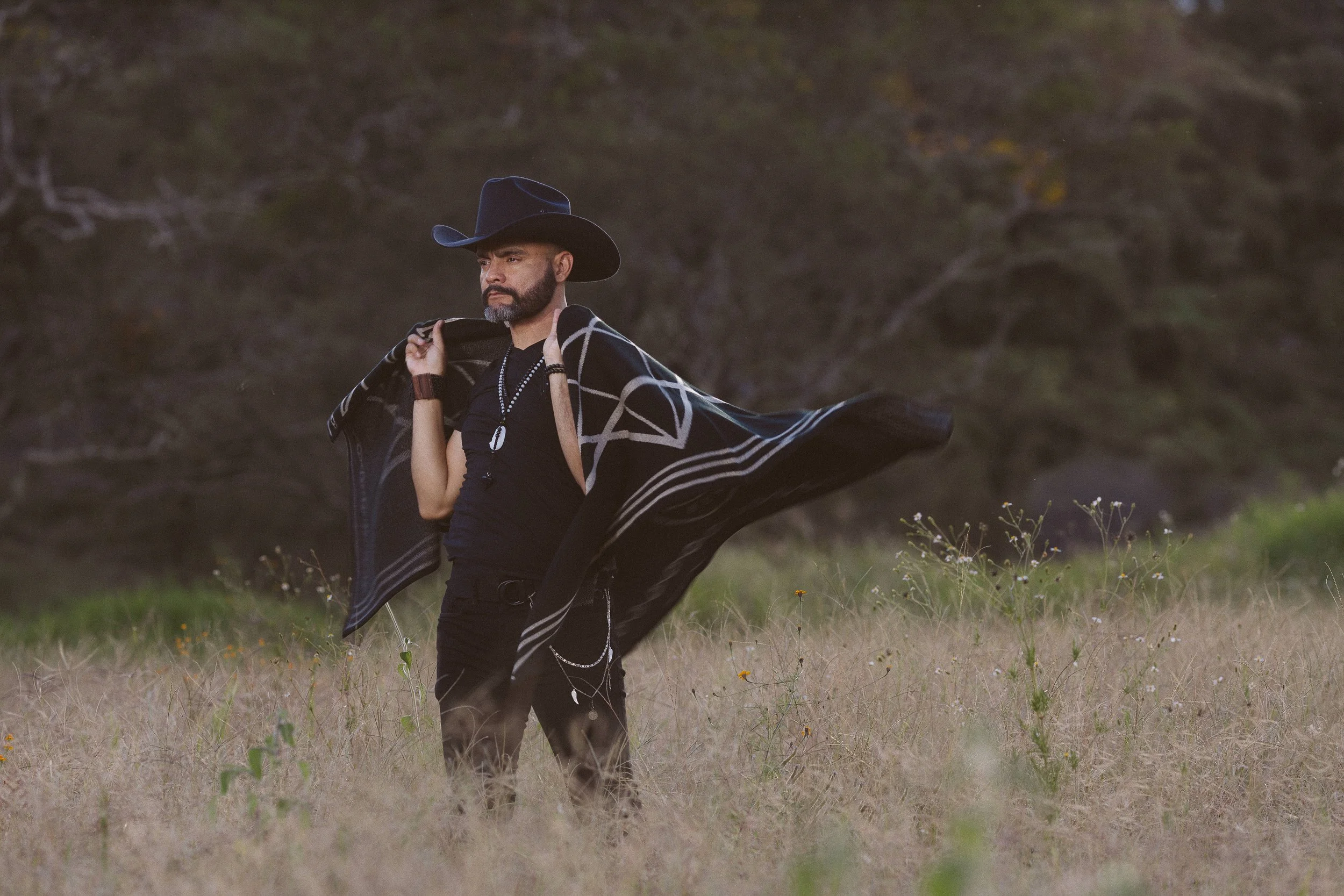 Hombre usando sombrero negro y poncho bordado, de pie en un campo rodeado de naturaleza.