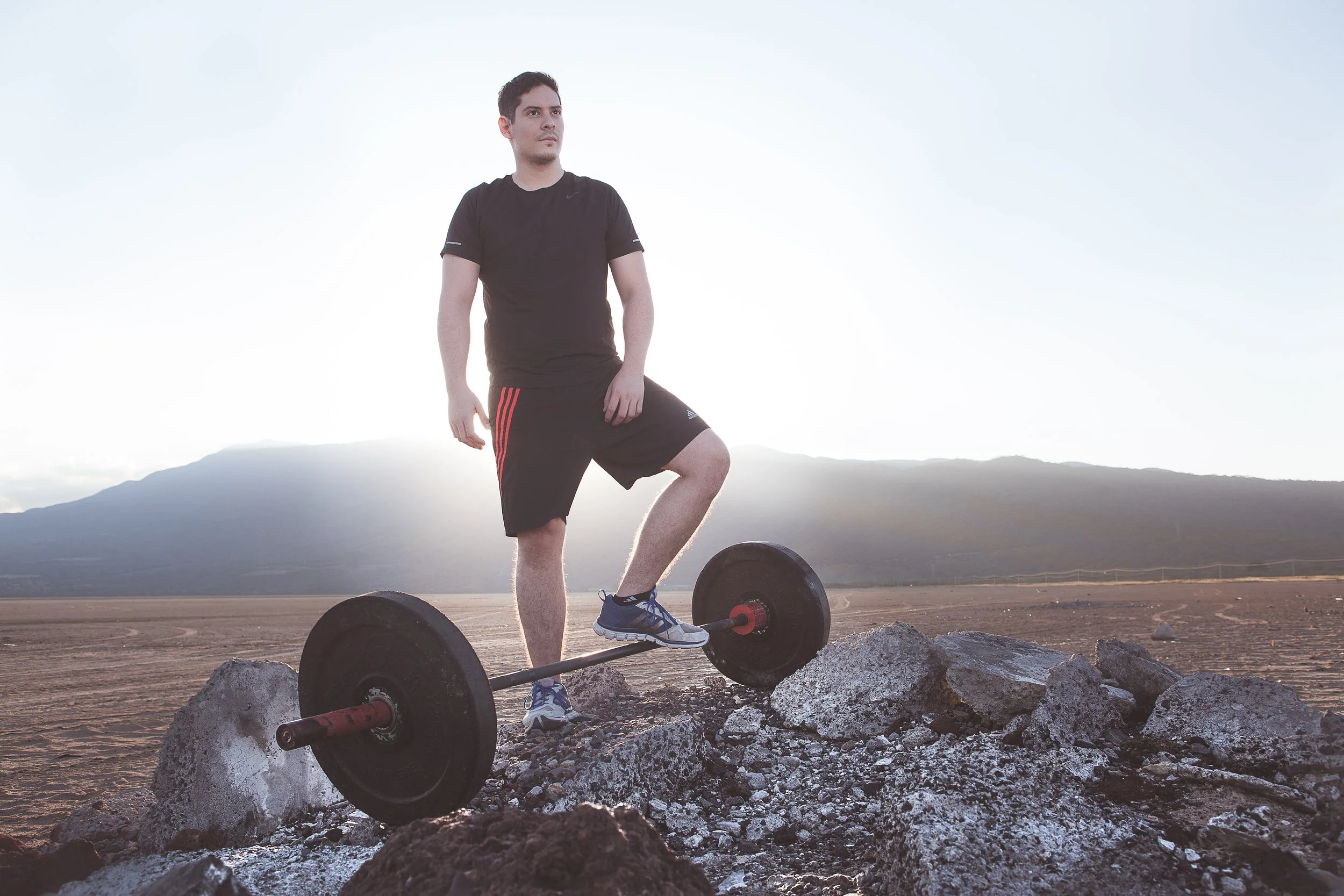 Hombre de pie sobre escombros con barra de pesas y paisaje montañoso al fondo.