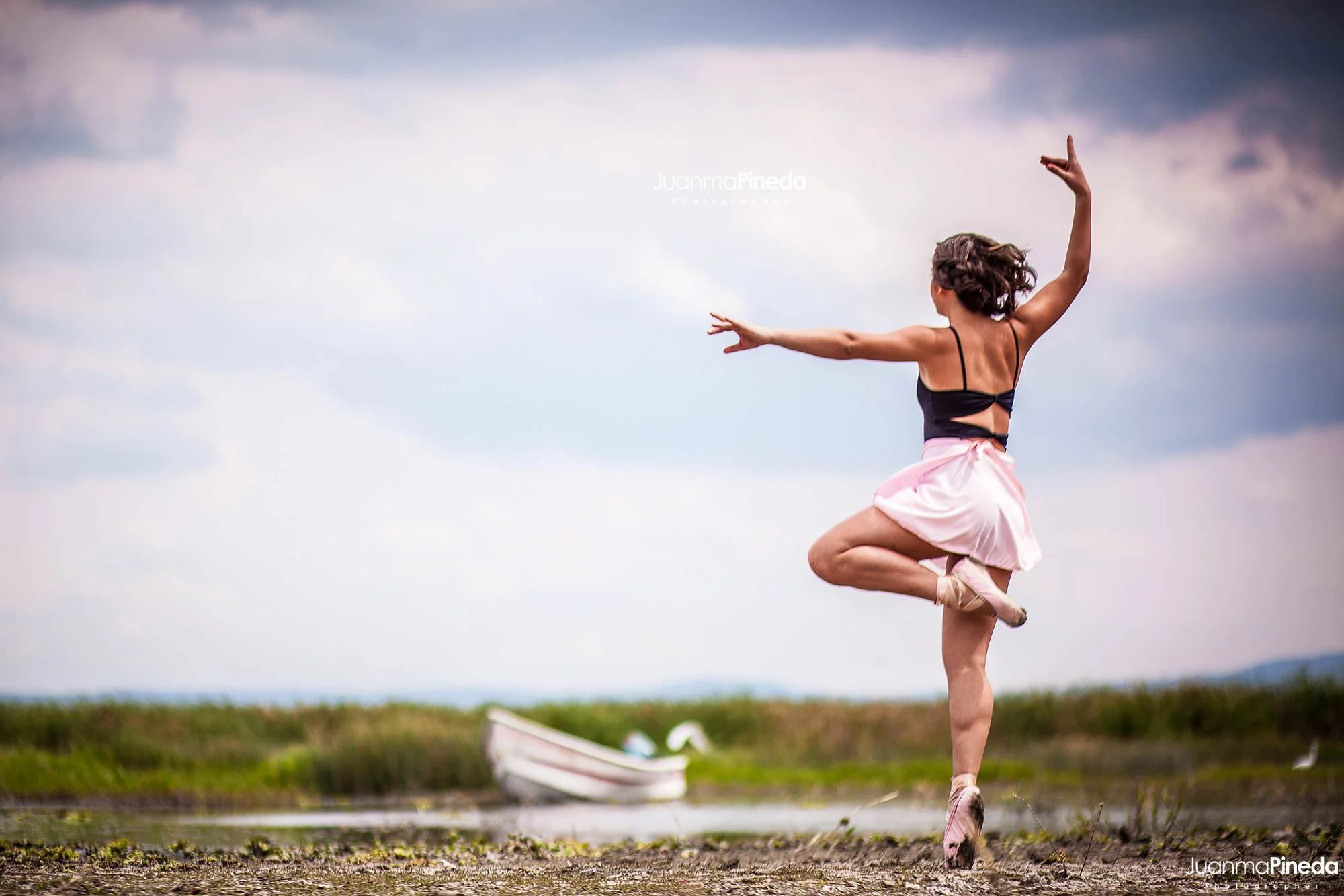 Bailarina en pose de ballet al aire libre cerca de un lago, con un bote al fondo.