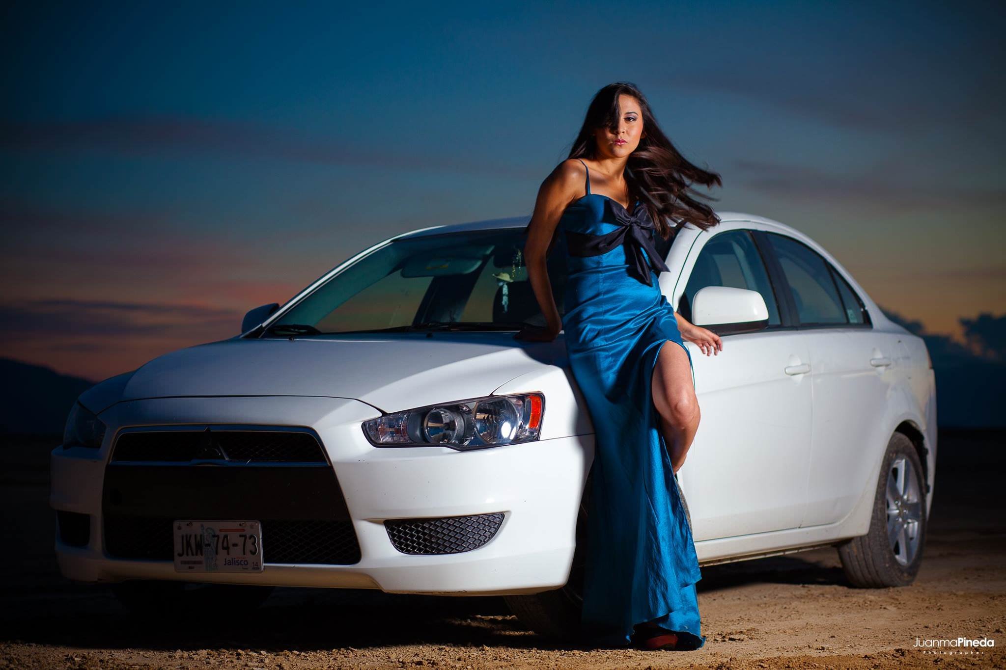 Mujer con vestido azul apoyada en un automóvil blanco al atardecer.