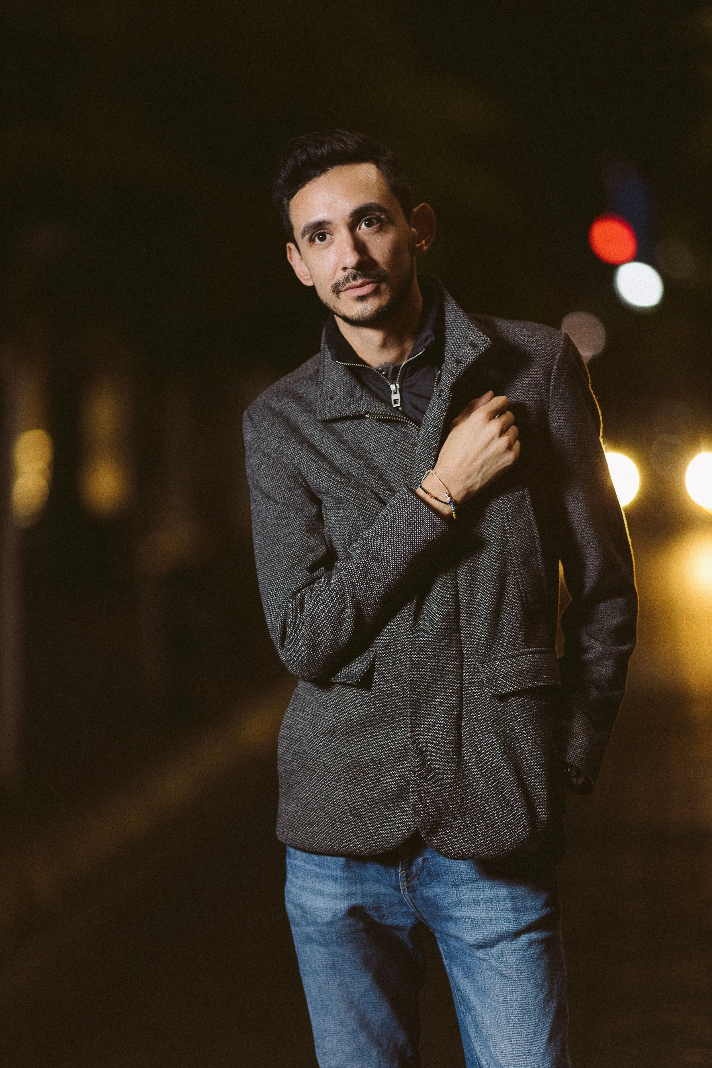 Hombre con chaqueta gris en la calle por la noche, luces difuminadas de fondo.