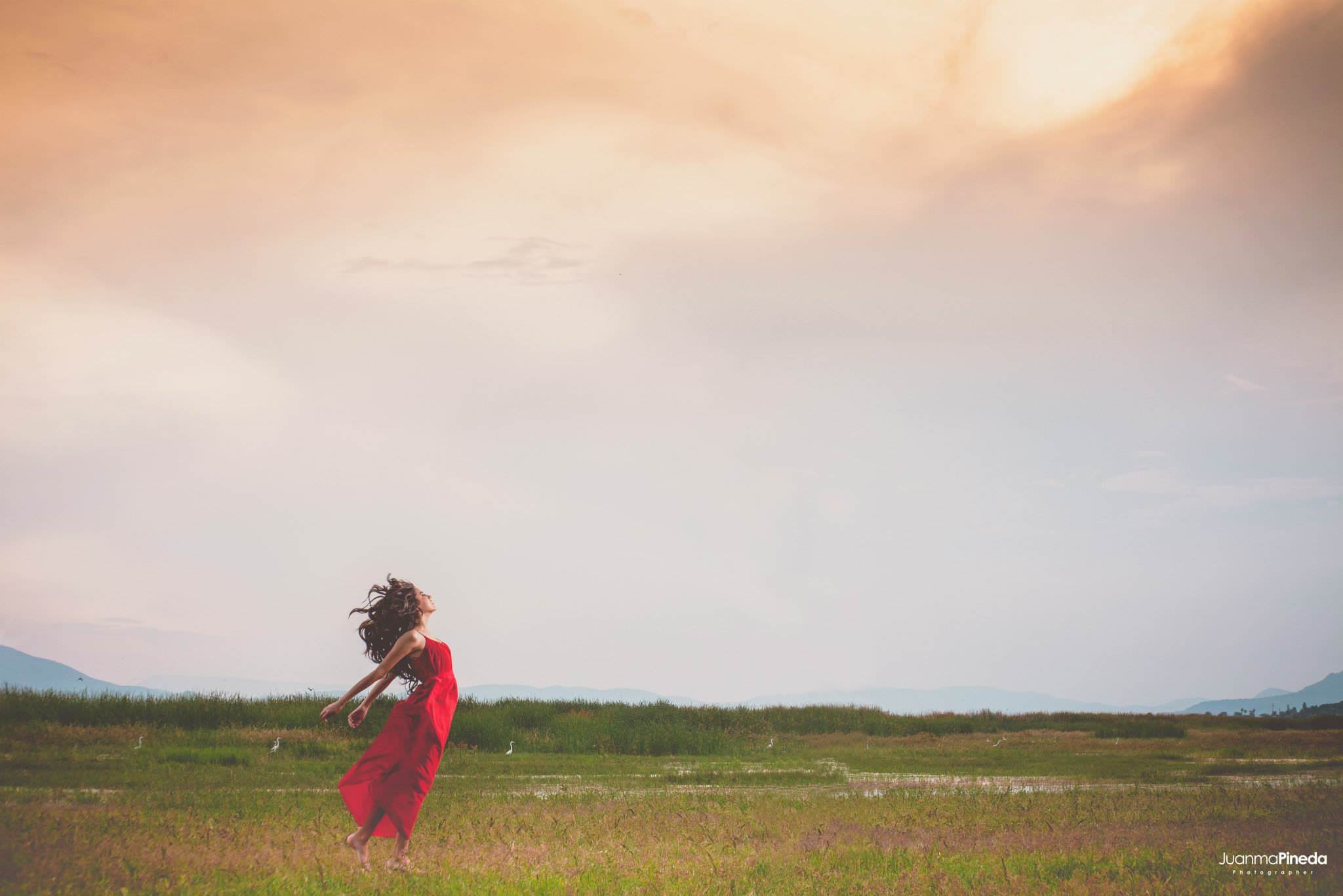 Mujer con vestido rojo en un campo con cielo nublado al atardecer.