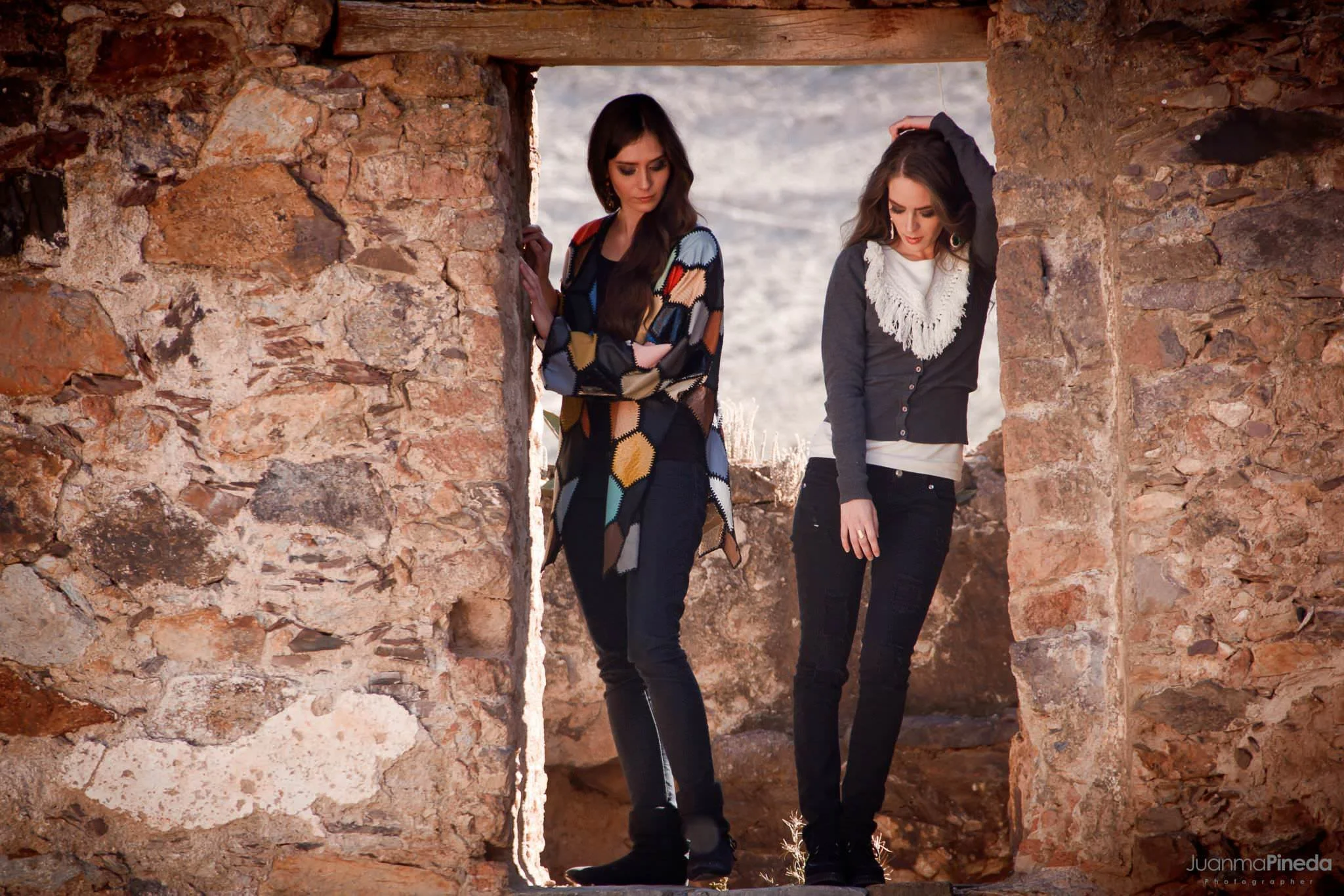 Dos mujeres paradas en un marco de piedra, ambas vestidas con ropa casual de invierno, en un entorno rústico al aire libre.