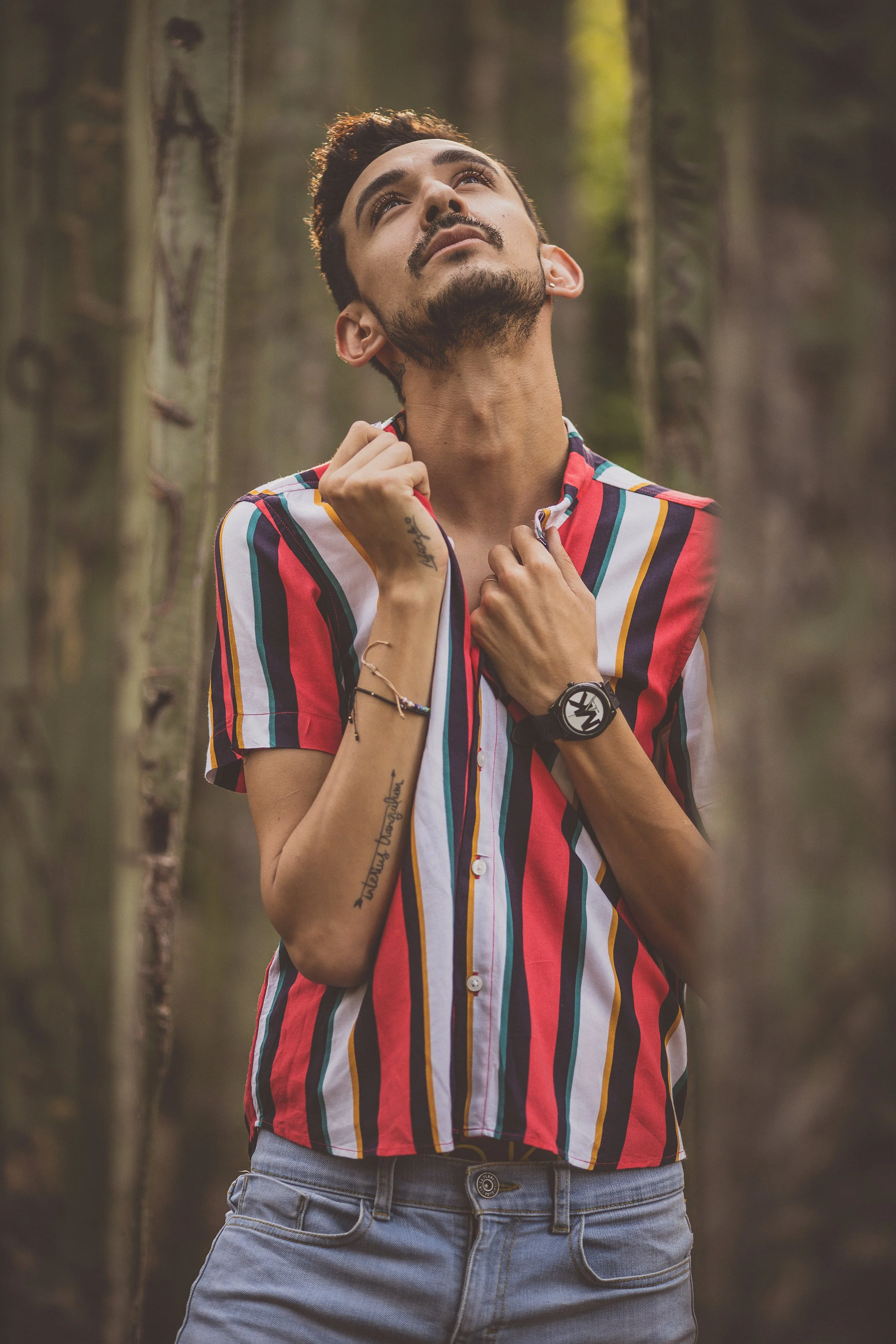 Persona con camisa de rayas coloridas en un entorno forestal, mirando hacia arriba con expresión reflexiva.