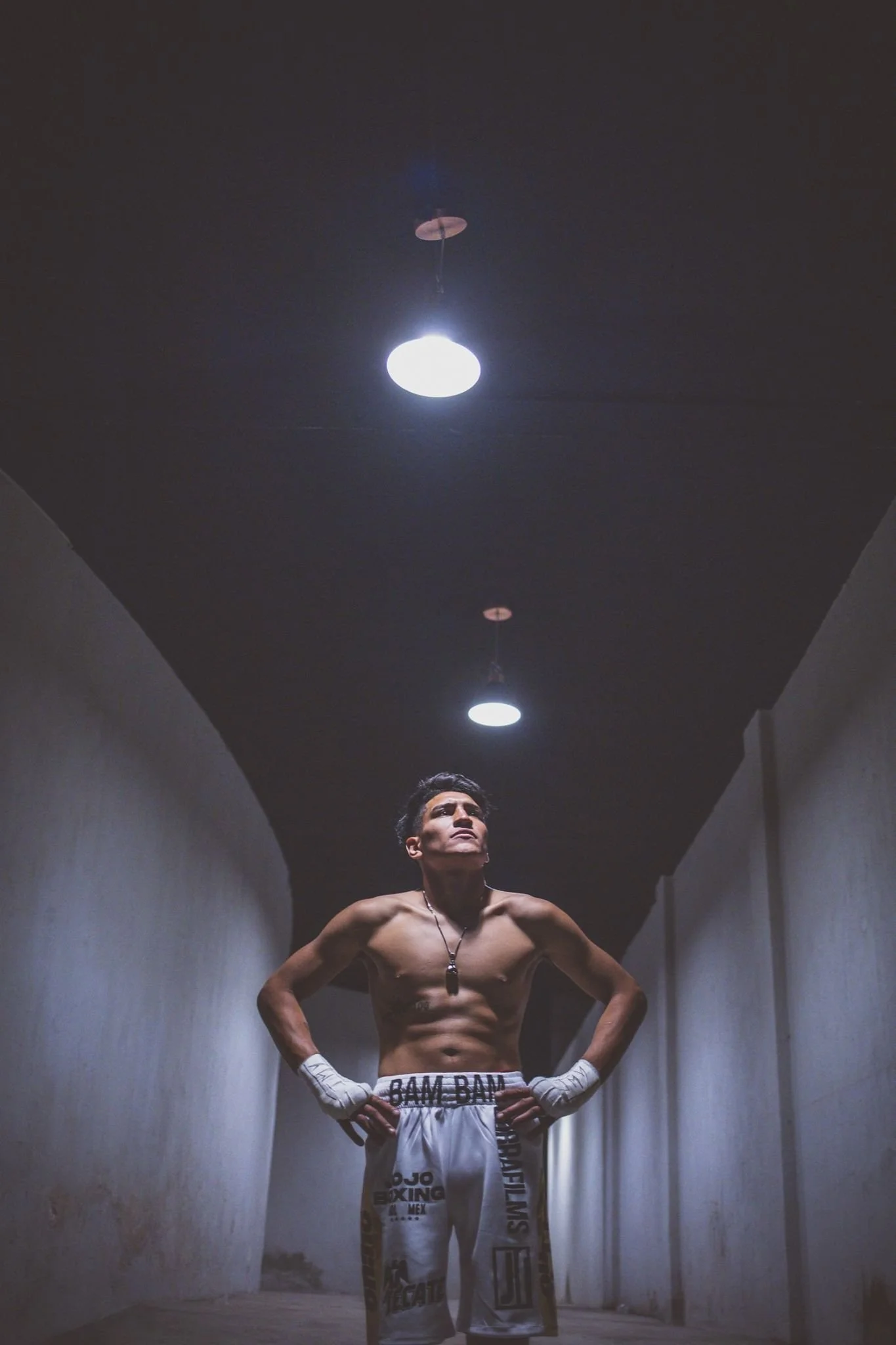 Boxeador sin camisa, usando pantalones cortos y vendaje en las manos, de pie en un pasillo oscuro con luces en el techo.