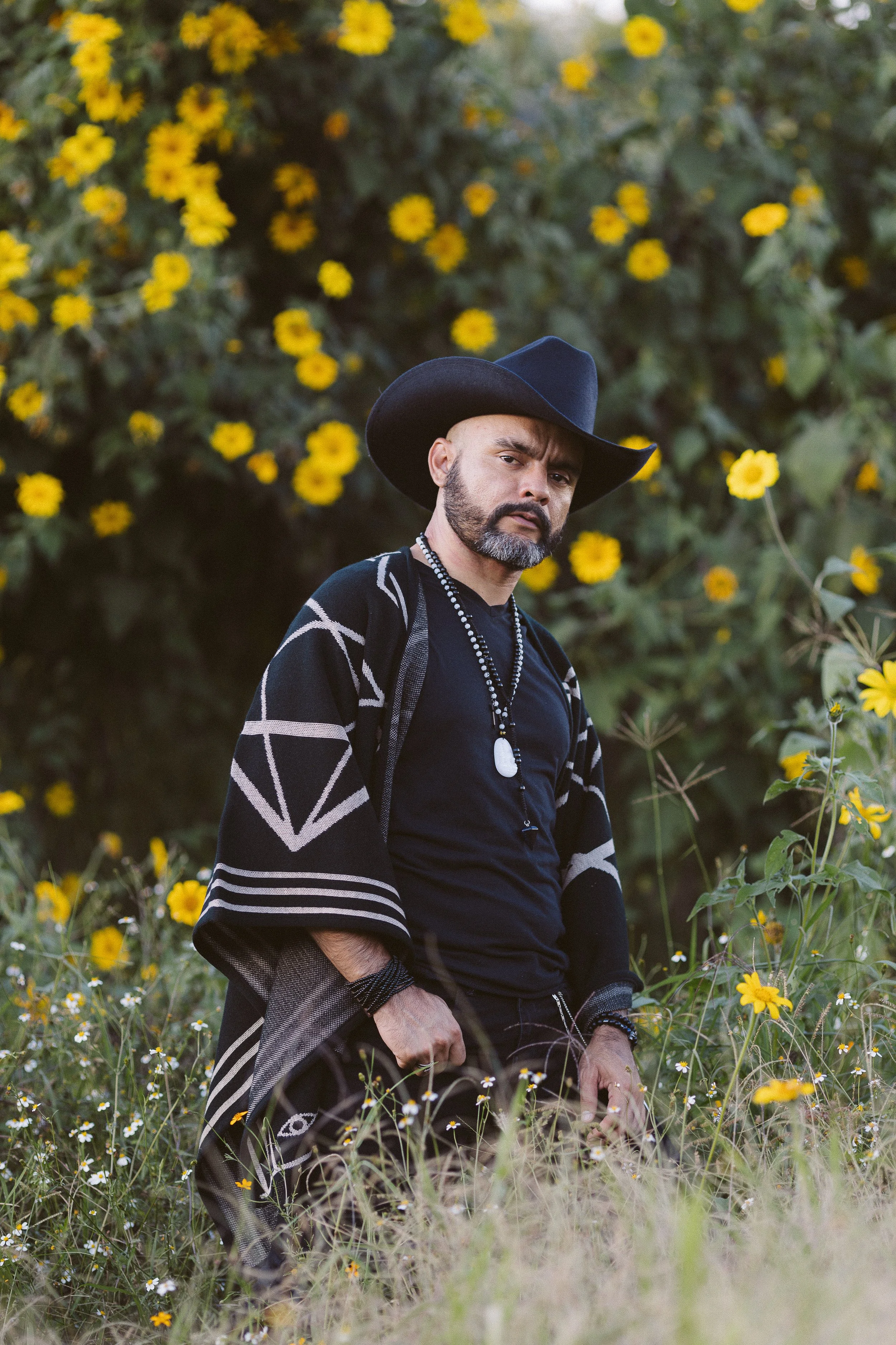 Hombre con sombrero y poncho de pie en un campo con flores amarillas.