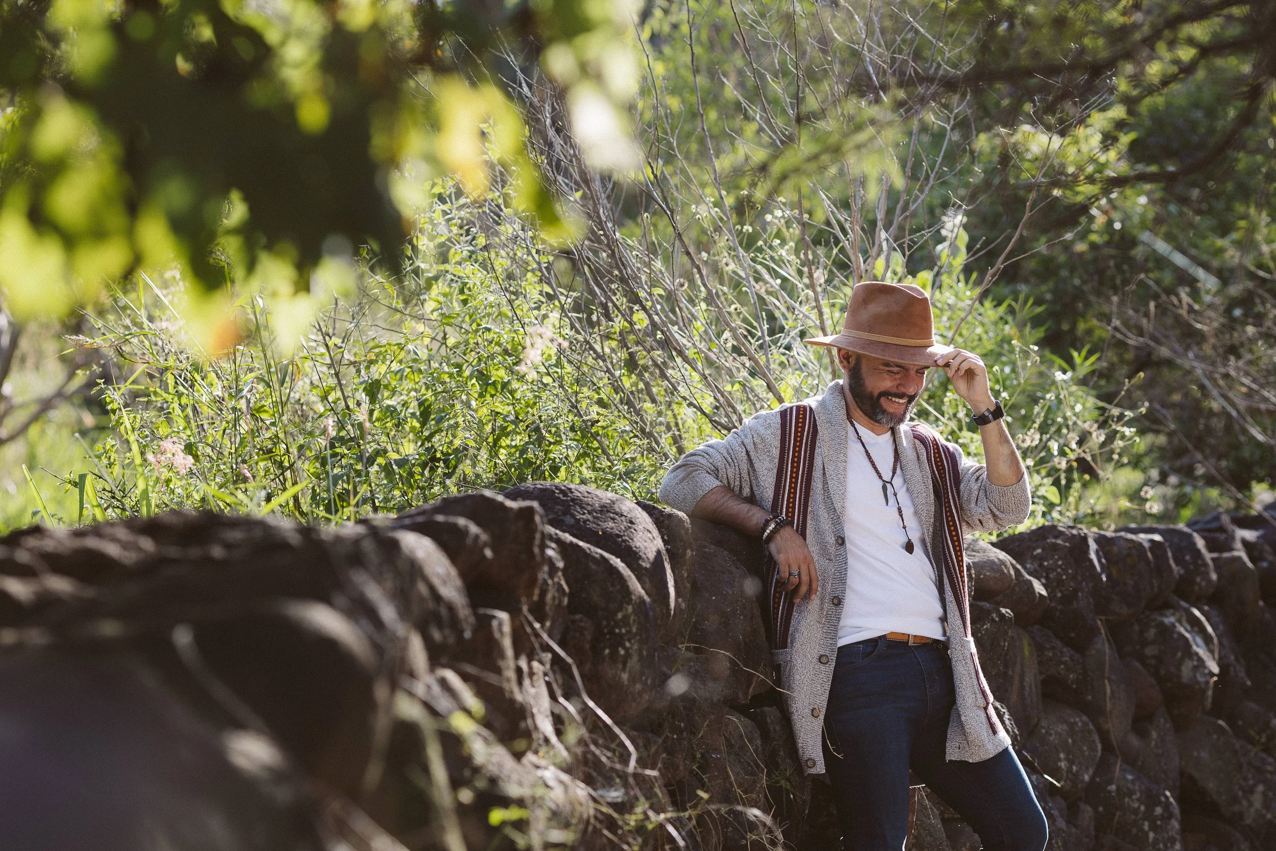 Hombre con sombrero posando al aire libre junto a un muro de piedra, rodeado de vegetación.