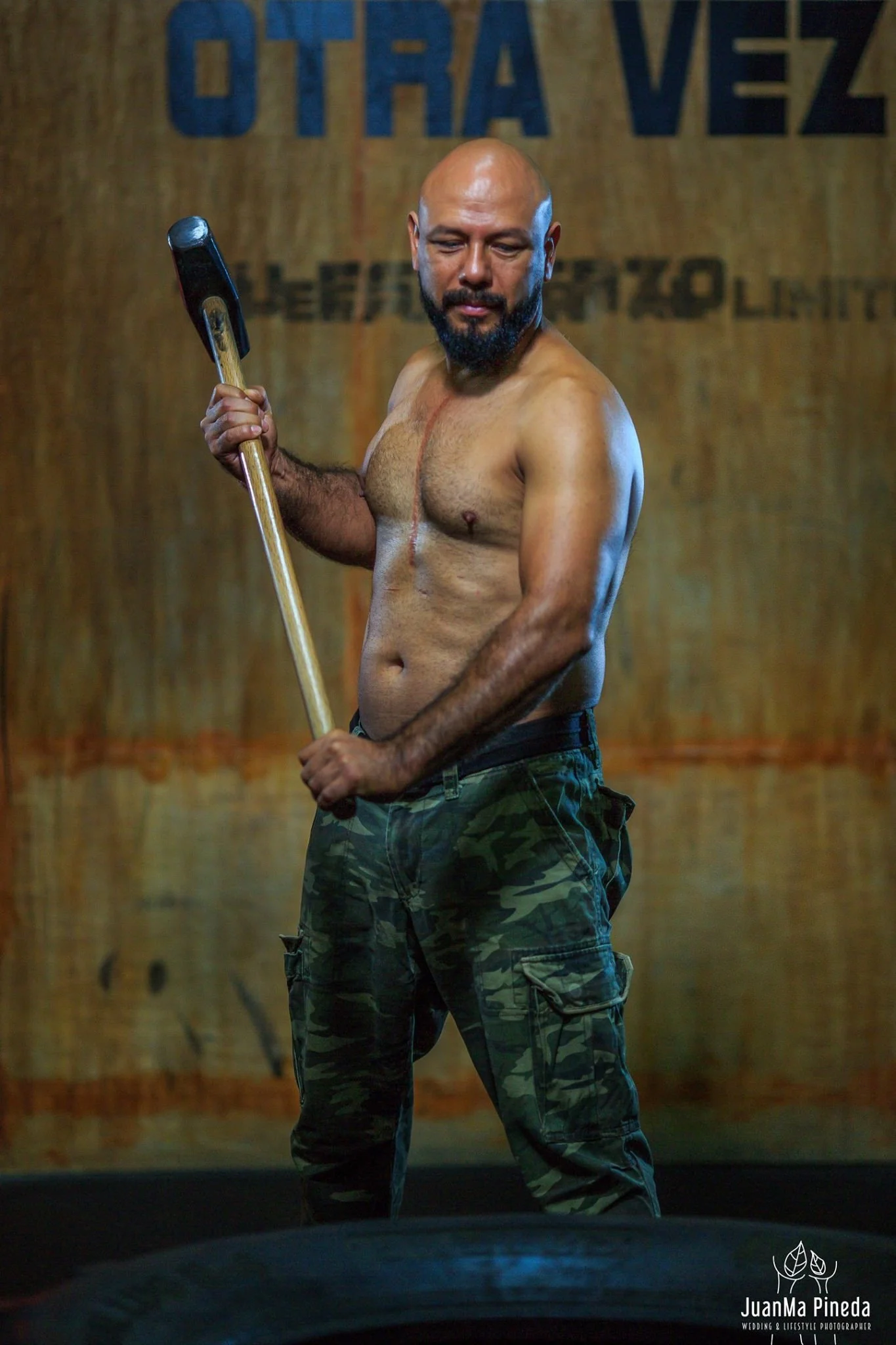 Hombre musculoso sin camisa, sosteniendo un mazo de manera firme. Viste pantalones camuflados y está frente a un fondo de madera con texto borroso.