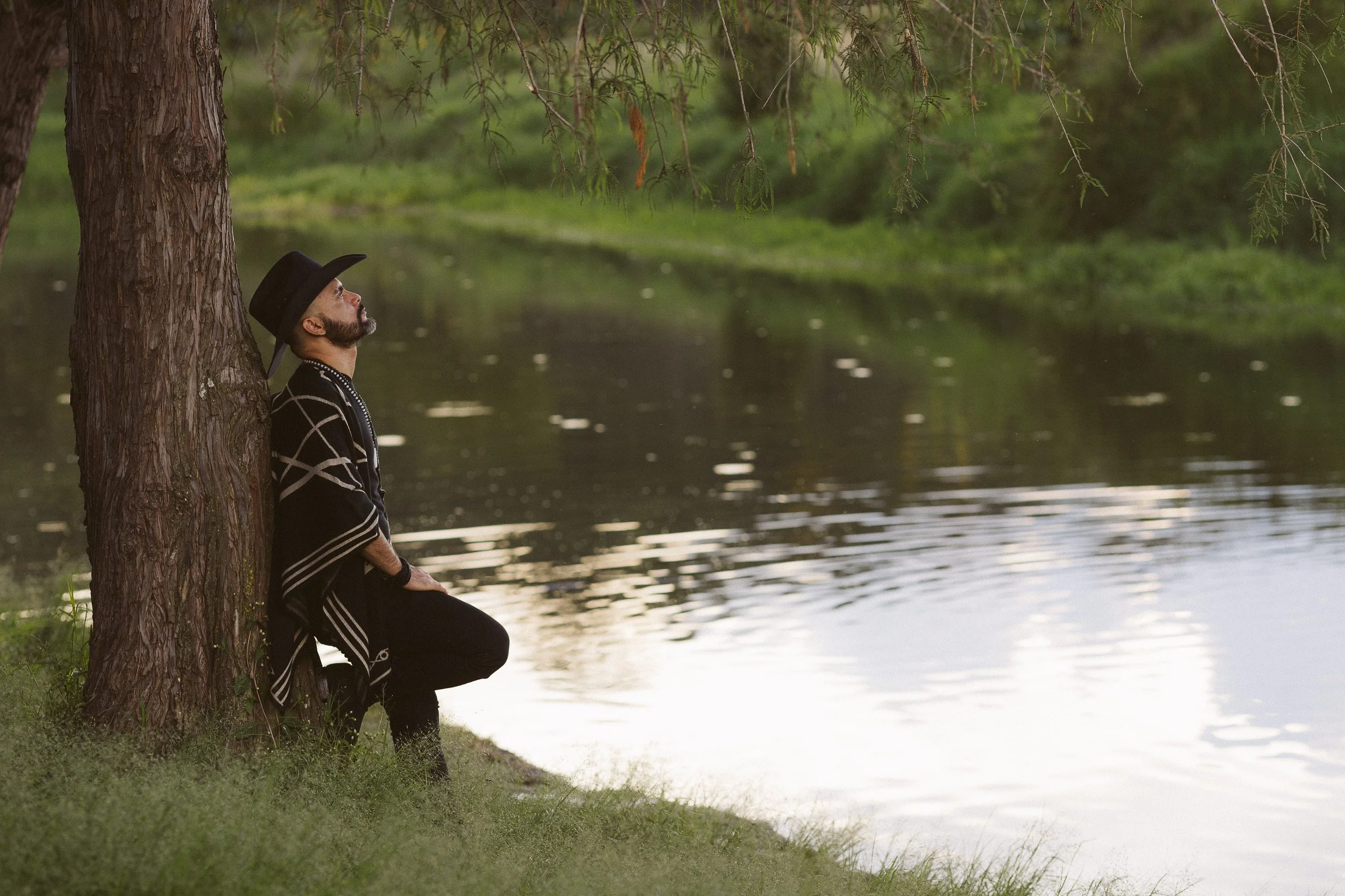 Hombre con sombrero y poncho descansando junto a un árbol al lado de un lago.