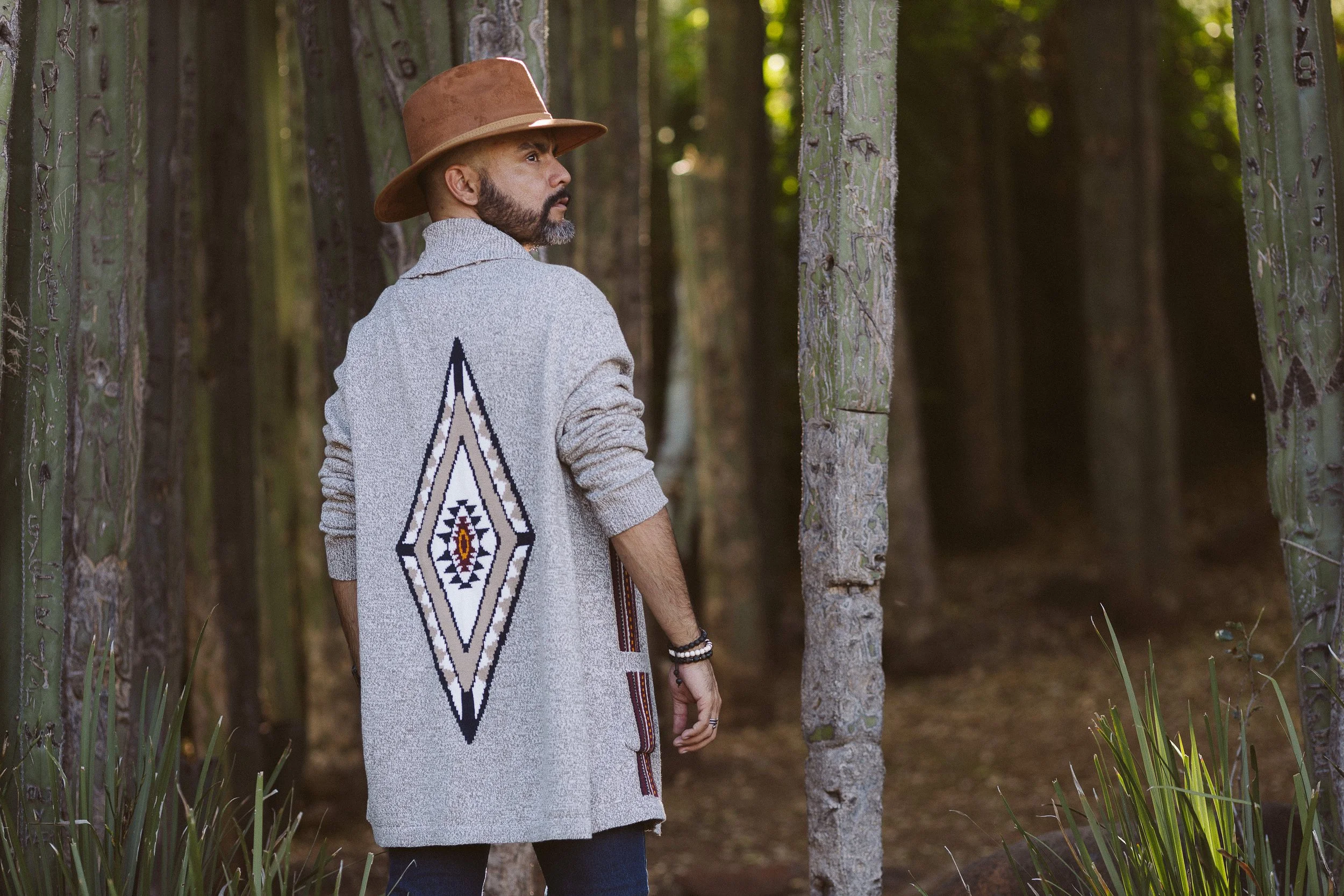 Hombre con sombrero marrón y suéter con diseño geométrico en bosque de cactus.