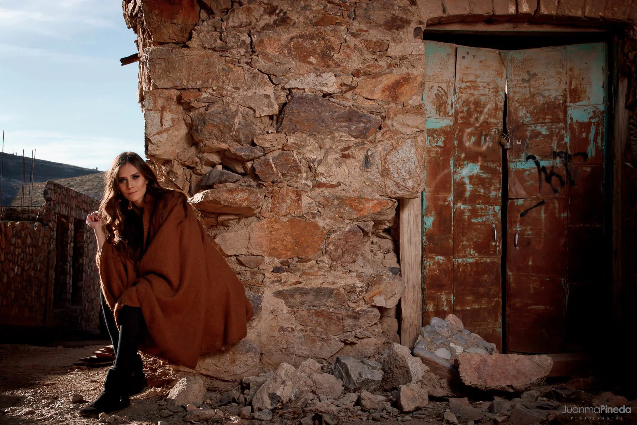 Mujer sentada en una construcción de piedra con una puerta antigua y paisaje montañoso al fondo.