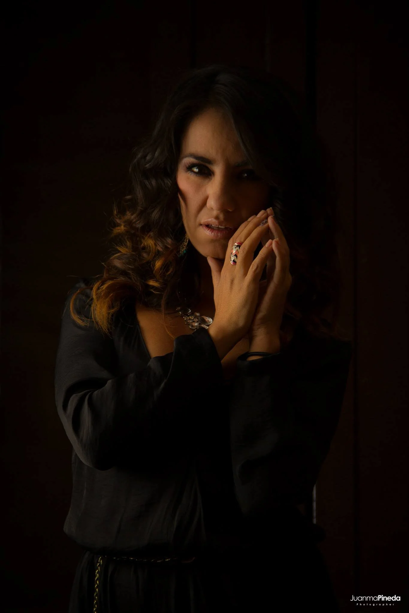 Mujer de cabello rizado y oscuro en fondo oscuro, vistiendo ropa negra y sosteniéndose la cara con las manos, lleva un anillo y collar.