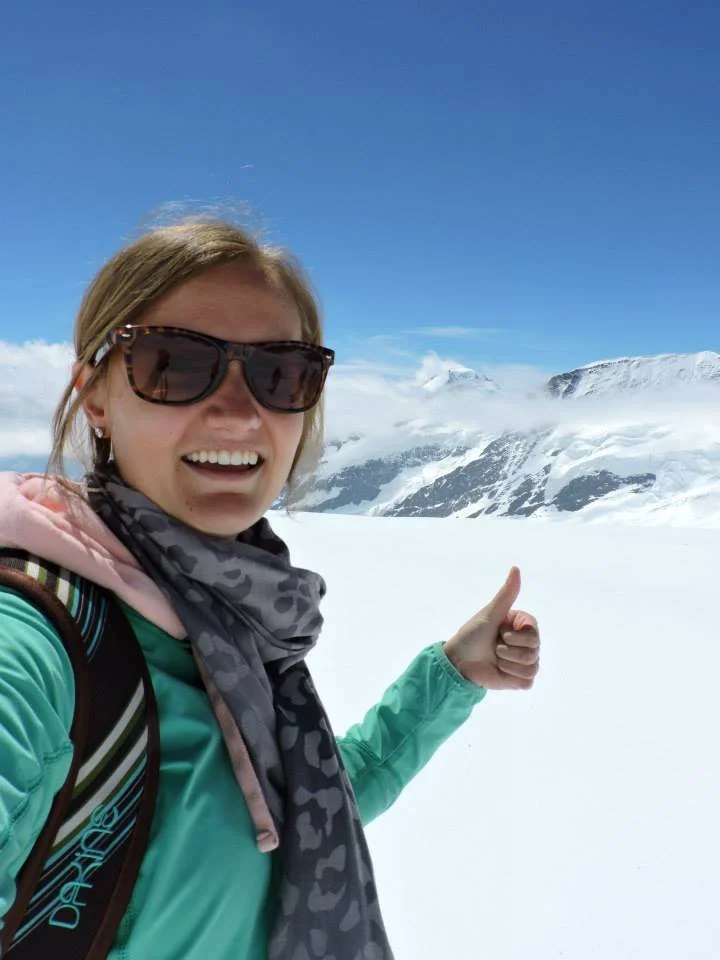 Woman in sunglasses and winter clothing smiling in a snowy mountain landscape, giving a thumbs-up.