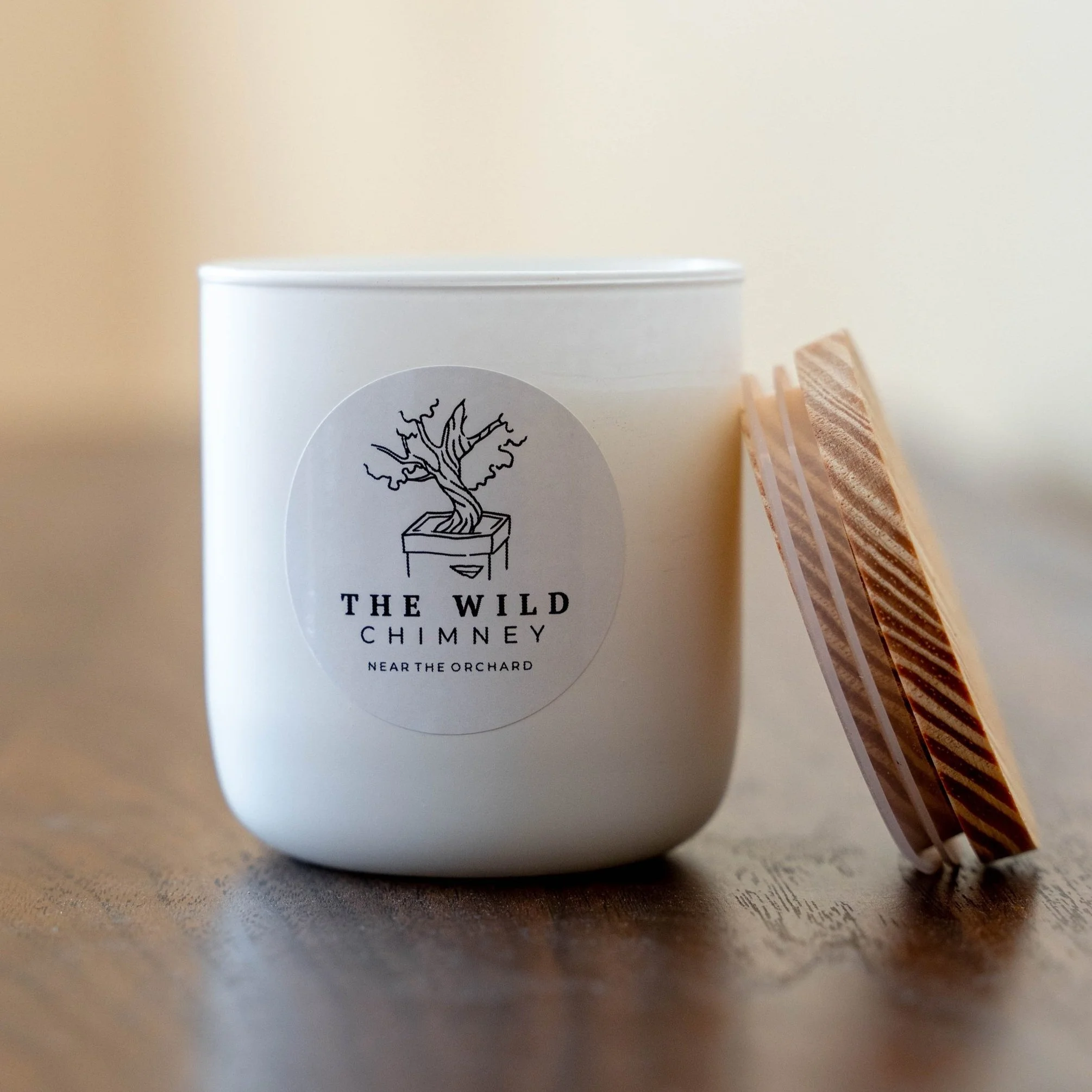 White candle in a container with a label showing a tree in a pot, labeled "The Wild Chimney, Near the Orchard." Next to the candle is a small stack of coasters or coiled paper pads on a wooden surface.