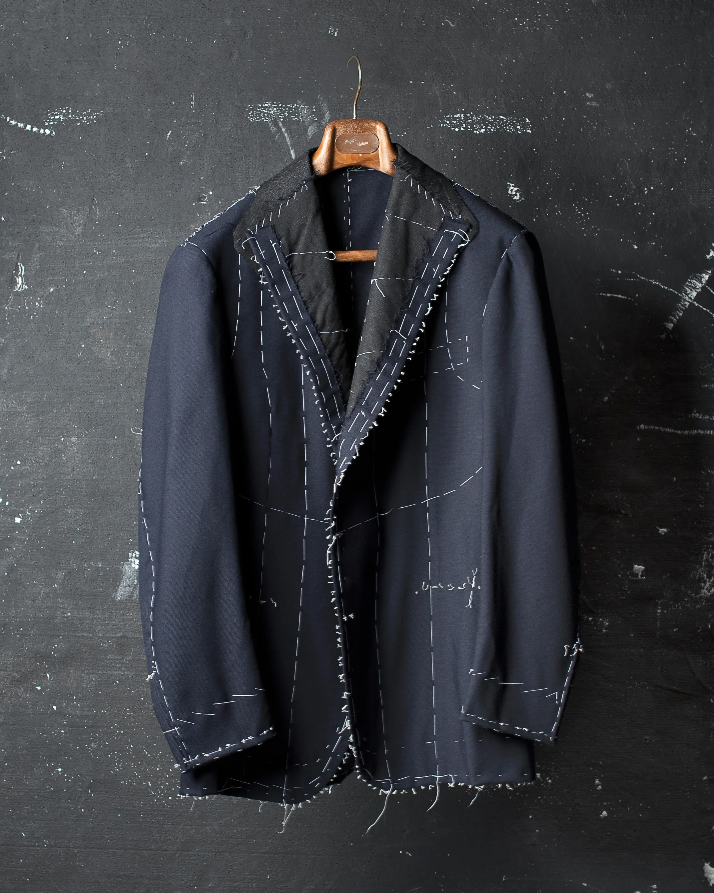 A black jacket with white stitching hang on a wooden hanger against a dark wall.