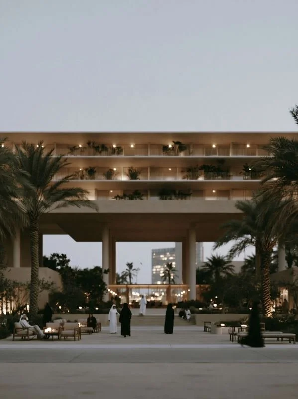 M4 · Árdh, concept image — a deep shaded colonnade under terraced floors at dusk, people sitting among palms