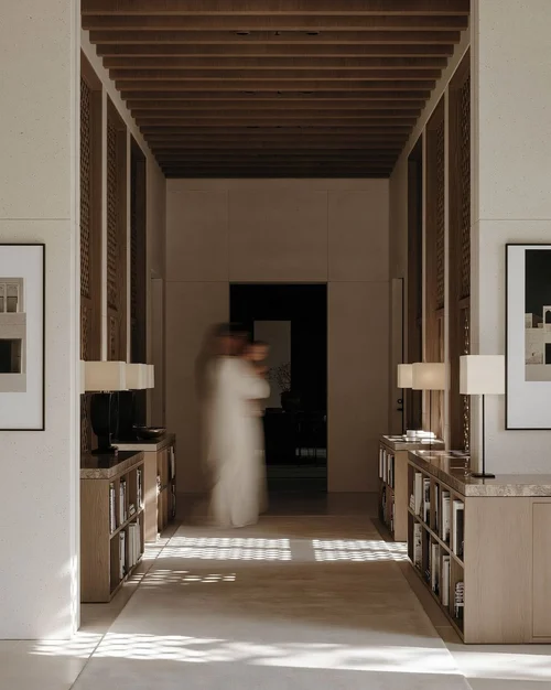 M18 · Árdh, concept image — a long timber-ceilinged corridor lined with low shelves and lamps, sunlight patterned on the floor, a figure in white walking through