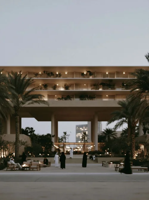 M4 · Árdh, concept image — a deep shaded colonnade under terraced floors at dusk, people sitting among palms